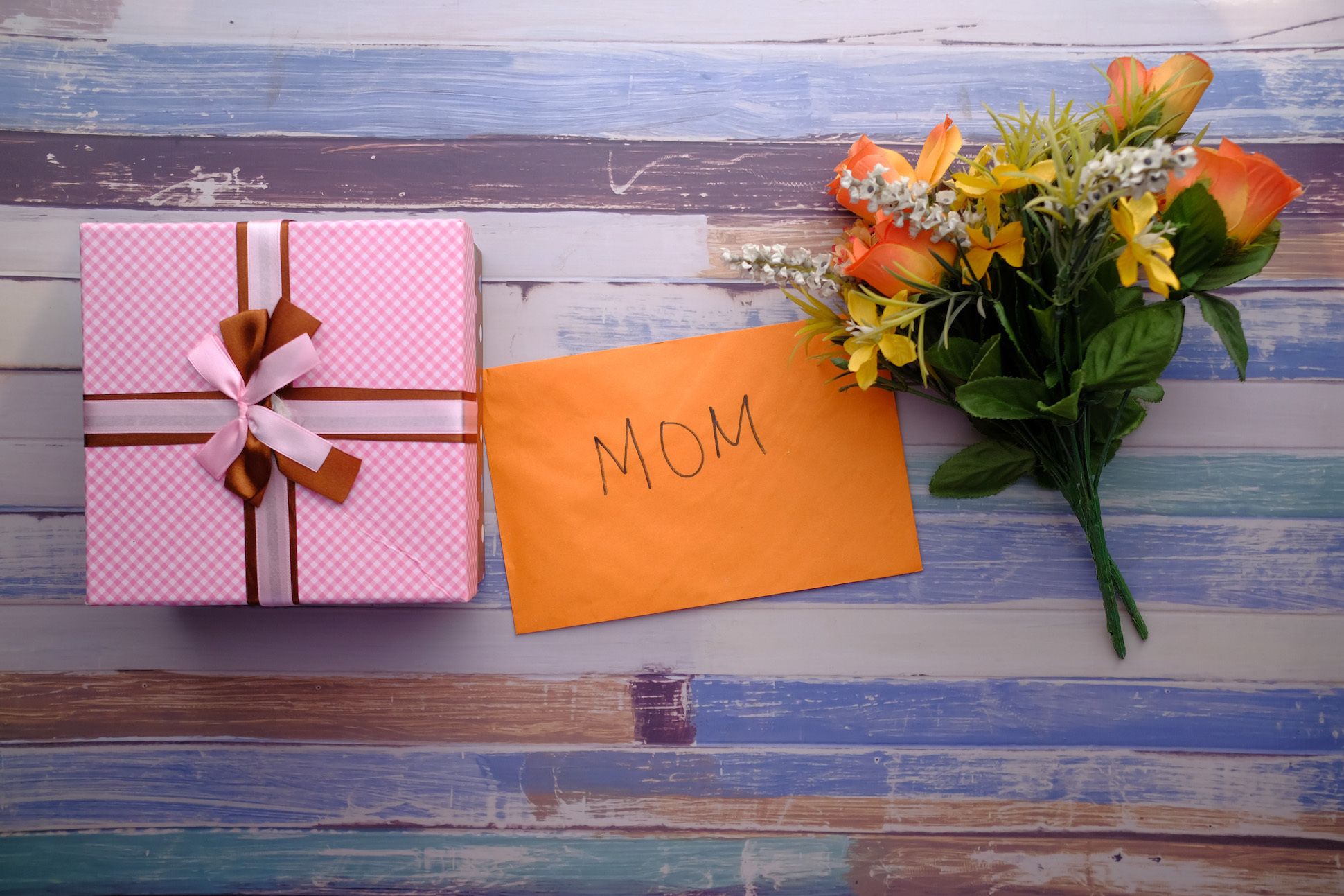 Image of a pink wrapped gift box, an orange envelop labeled MOM, and a yellow/orange/white floral arrangement.