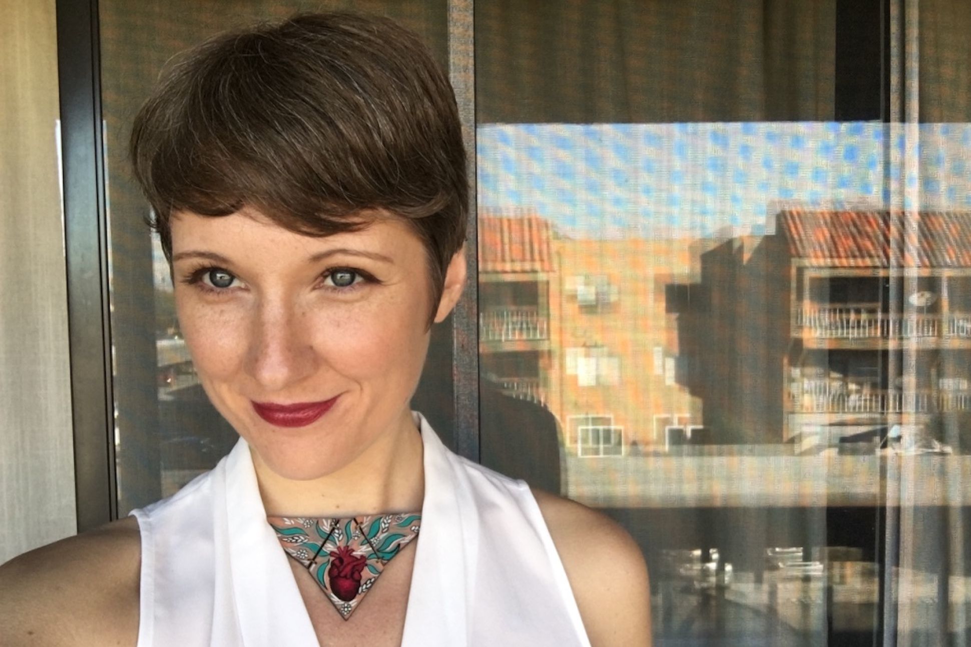 A short haired woman wearing a white vest with a rose neck piece in front of a balcony door