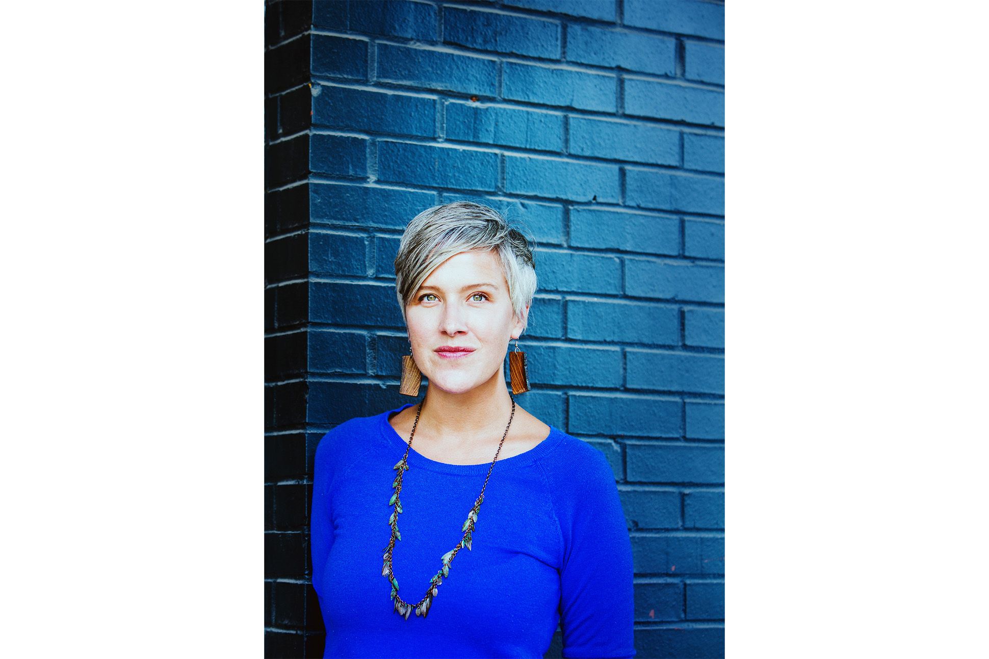 Headshot of poet Kristi Maxwell wearing a blue long sleeve blouse and a necklace in front of a blue painted wall