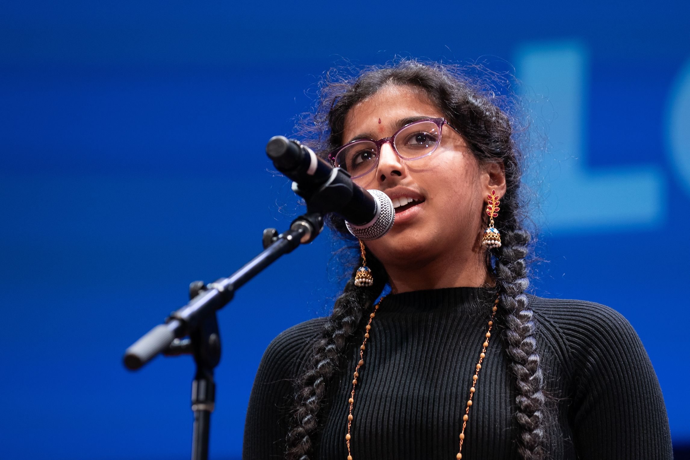 2023 Poetry Out Loud National Champion Sreepadaarchana Munjuluri, a teen girl wearing glasses, vibrant earrings, and a copper necklace with a black sweater. Her black hair is in two braids and she is standing at a microphone.