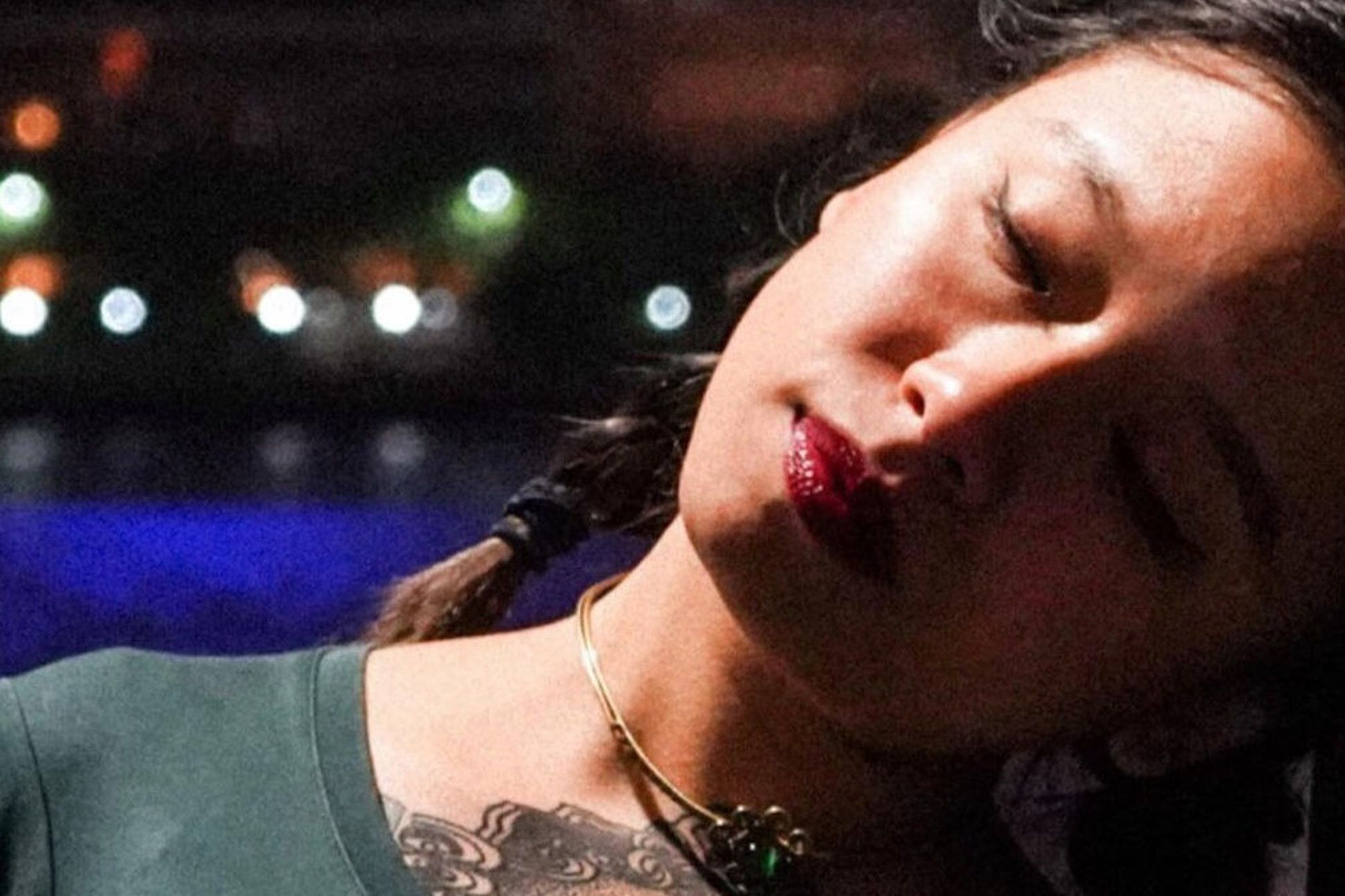 An Asian woman wearing a choker and with tattoos are her chest and tilting her head