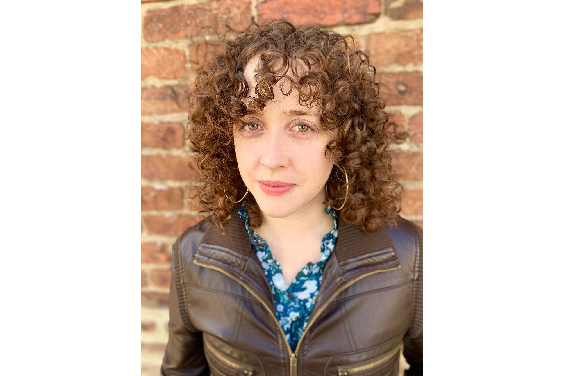 A white woman with curly hair in a leather jacket standing in front of a brick wall