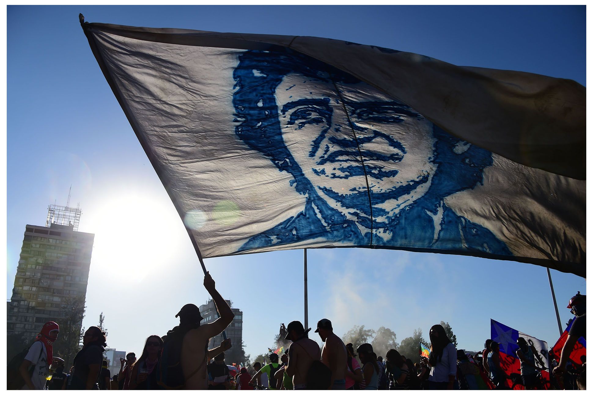 Photo of protestors in Santiago, one protestor holding large white flag with image of Jara's face, against blue sky.