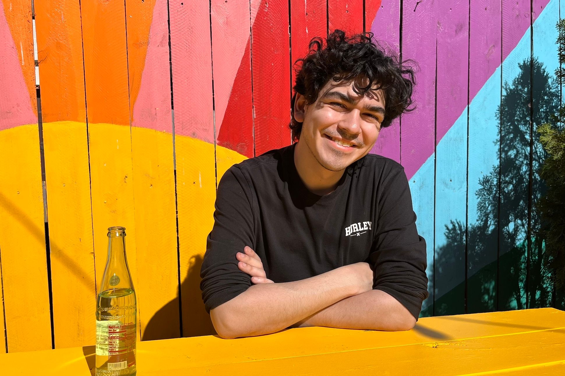 Julián Martinez, a Latino man with
curly dark hair, sits at a picnic bench in front of a rainbow-painted fence, a
glass bottle of water at his side.