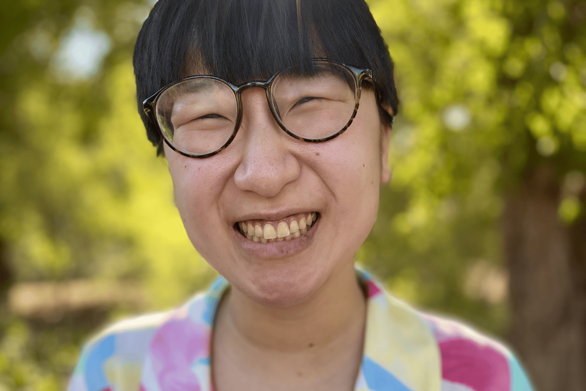 A short hair, spectacled Asian woman wearing a colorful shirt and smiling in front of trees