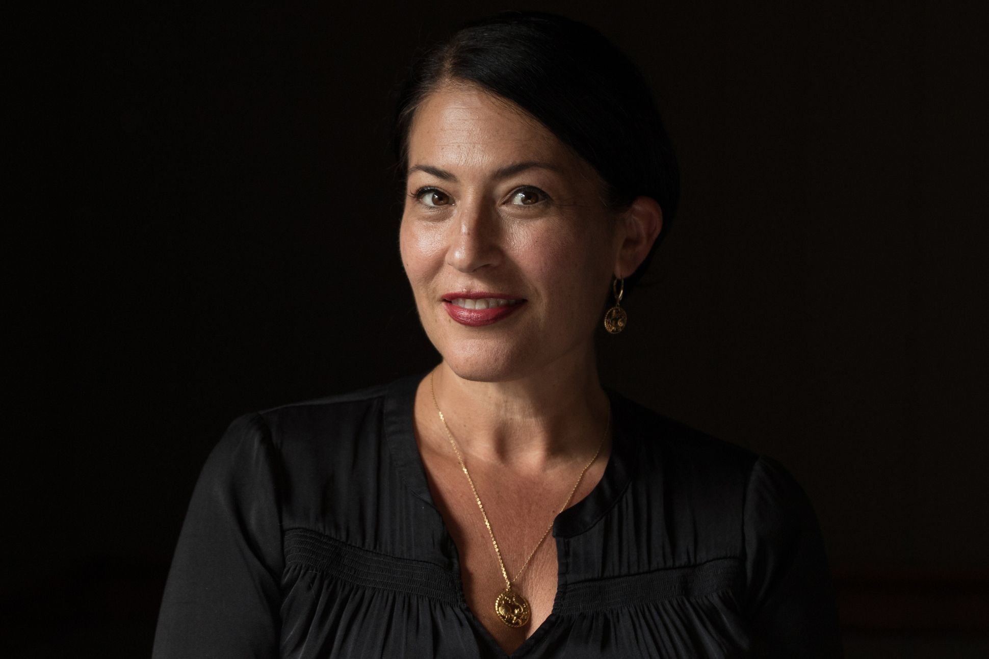 A light/medium-skinned woman with straight dark hair parted to the side and pulled back (Ada Limón) smiles softly. She is wearing a berry lip color, a long-sleeved black top, a gold pendant necklace, and gold earrings that are slightly obscured in the bla