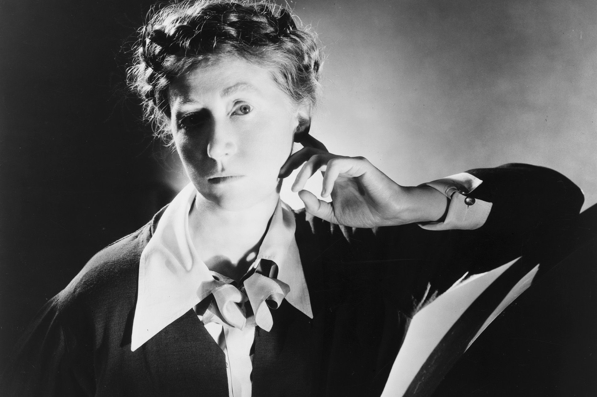Marianne Moore in a button down shirt, with her left hand touching her ear.