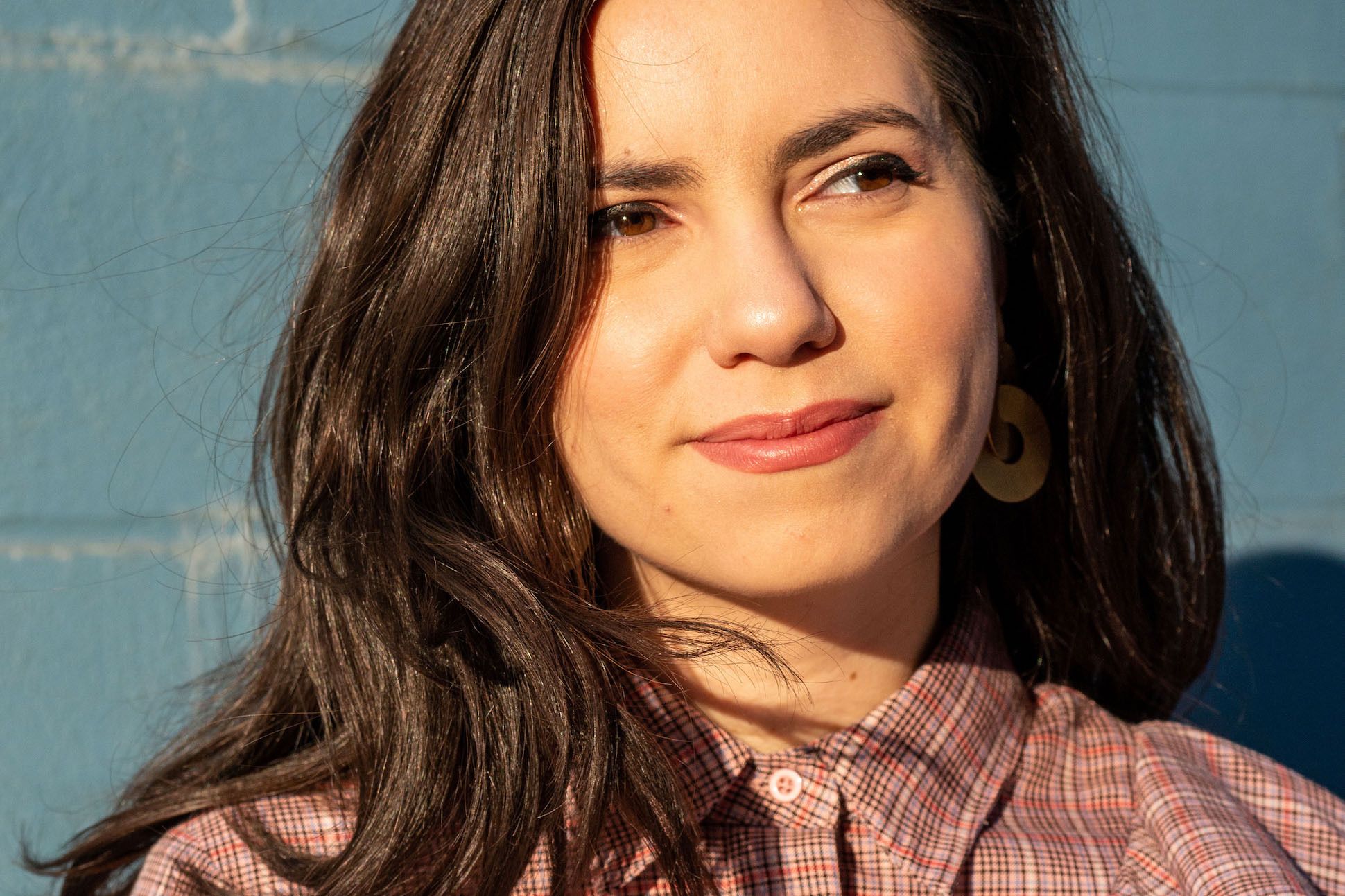 A long haired woman in a plaid shirt in front of a teal colored brick wall