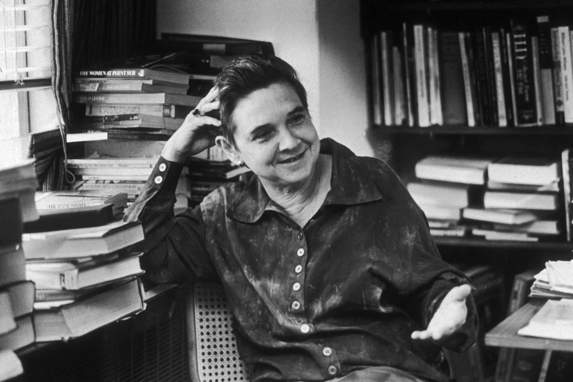 Black and white photograph of Adrienne Rich sitting at a desk, surrounded by piles of books.