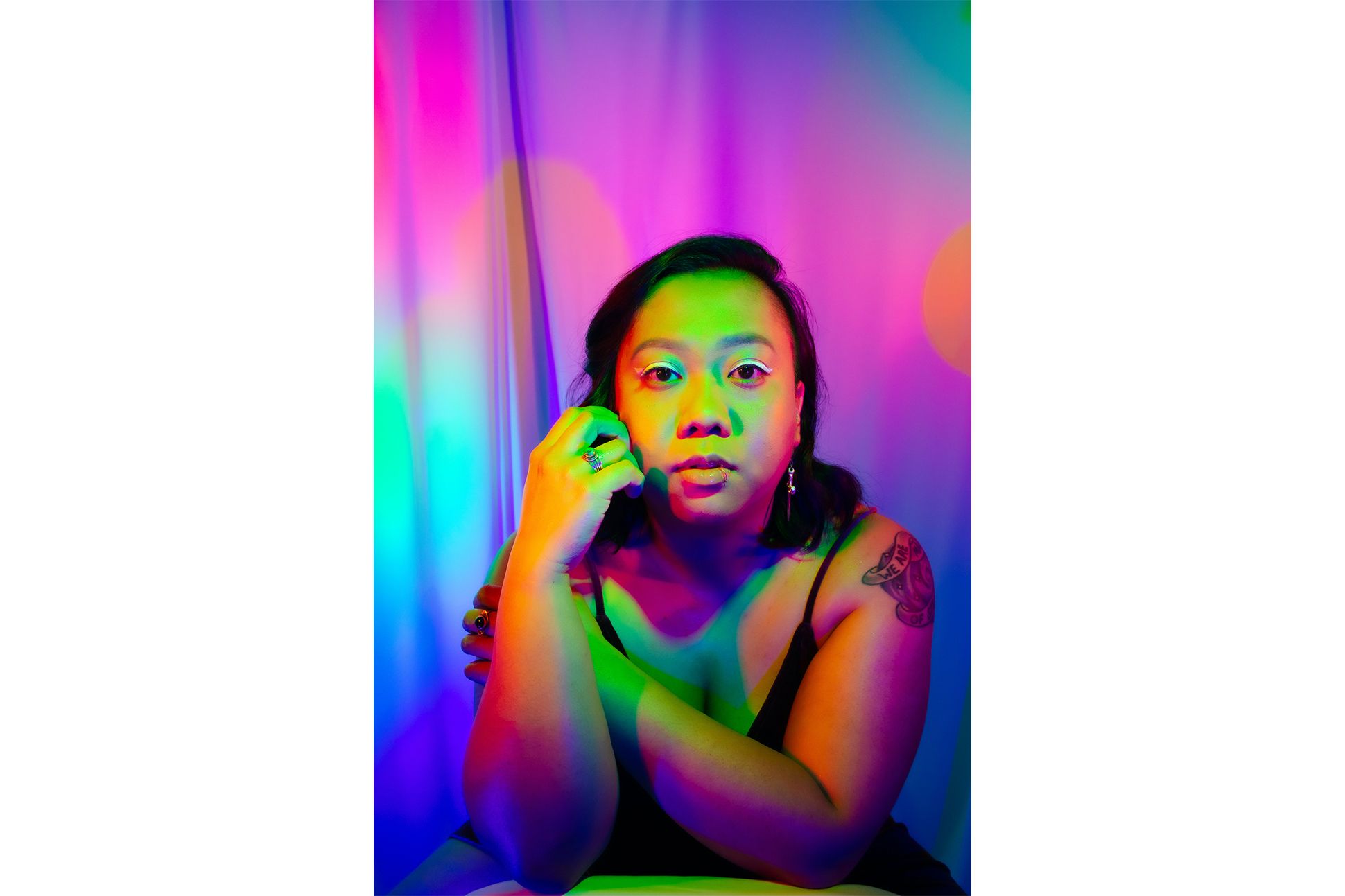 Filipinx American poet Cariño Ina looking at the camera with her hand on her face in a black cami top under colorful neon lights