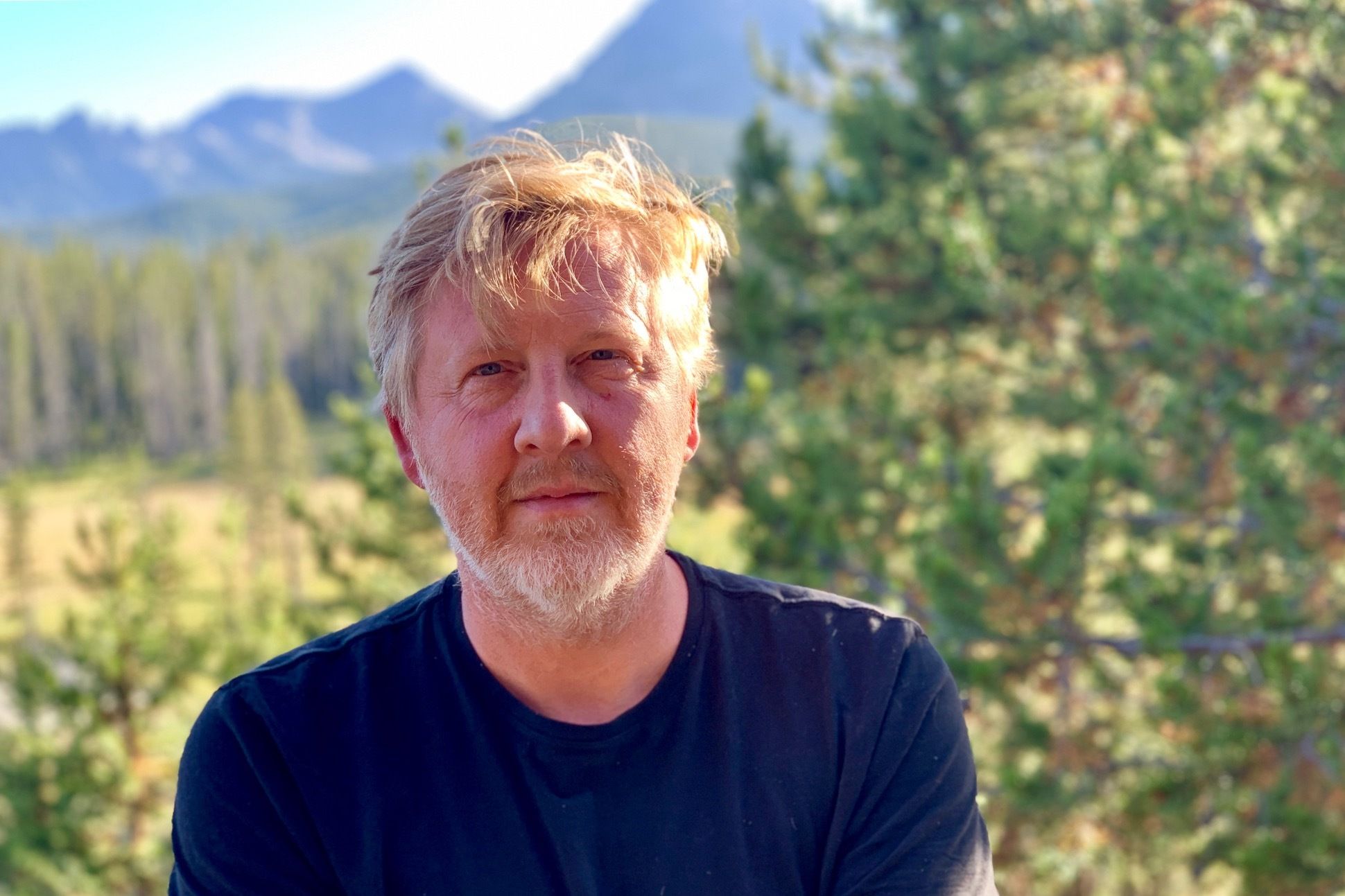 A man with blond hair and beard. The background has a forest and mountains.