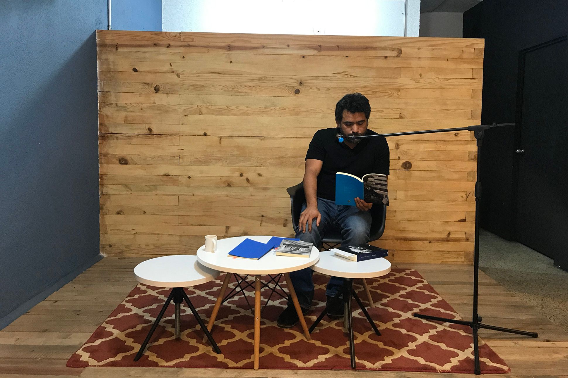 A man sits behind three nested tables reading from a book into a microphone