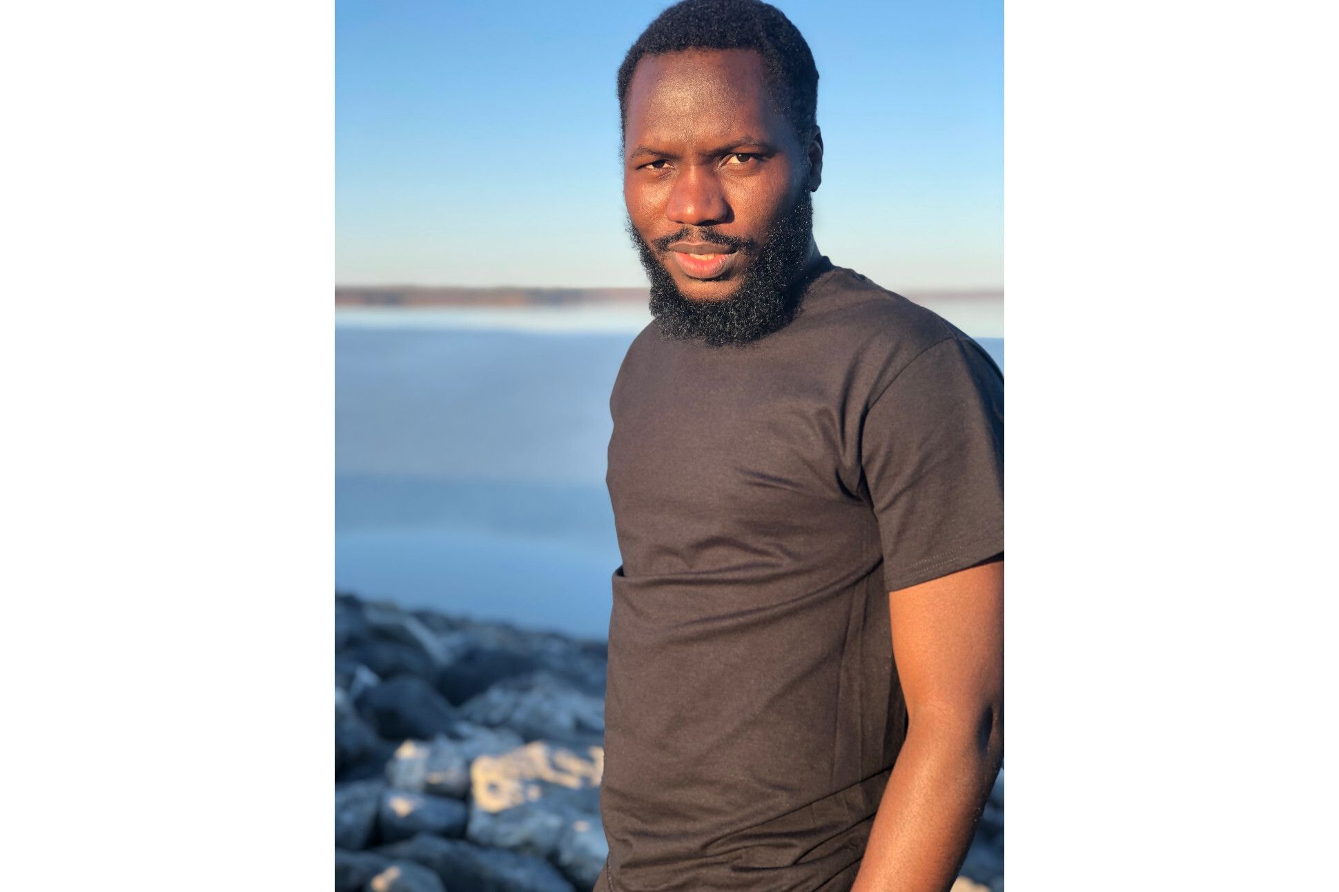 A black man in a grey t-shirt standing by a beach under a clear blue sky