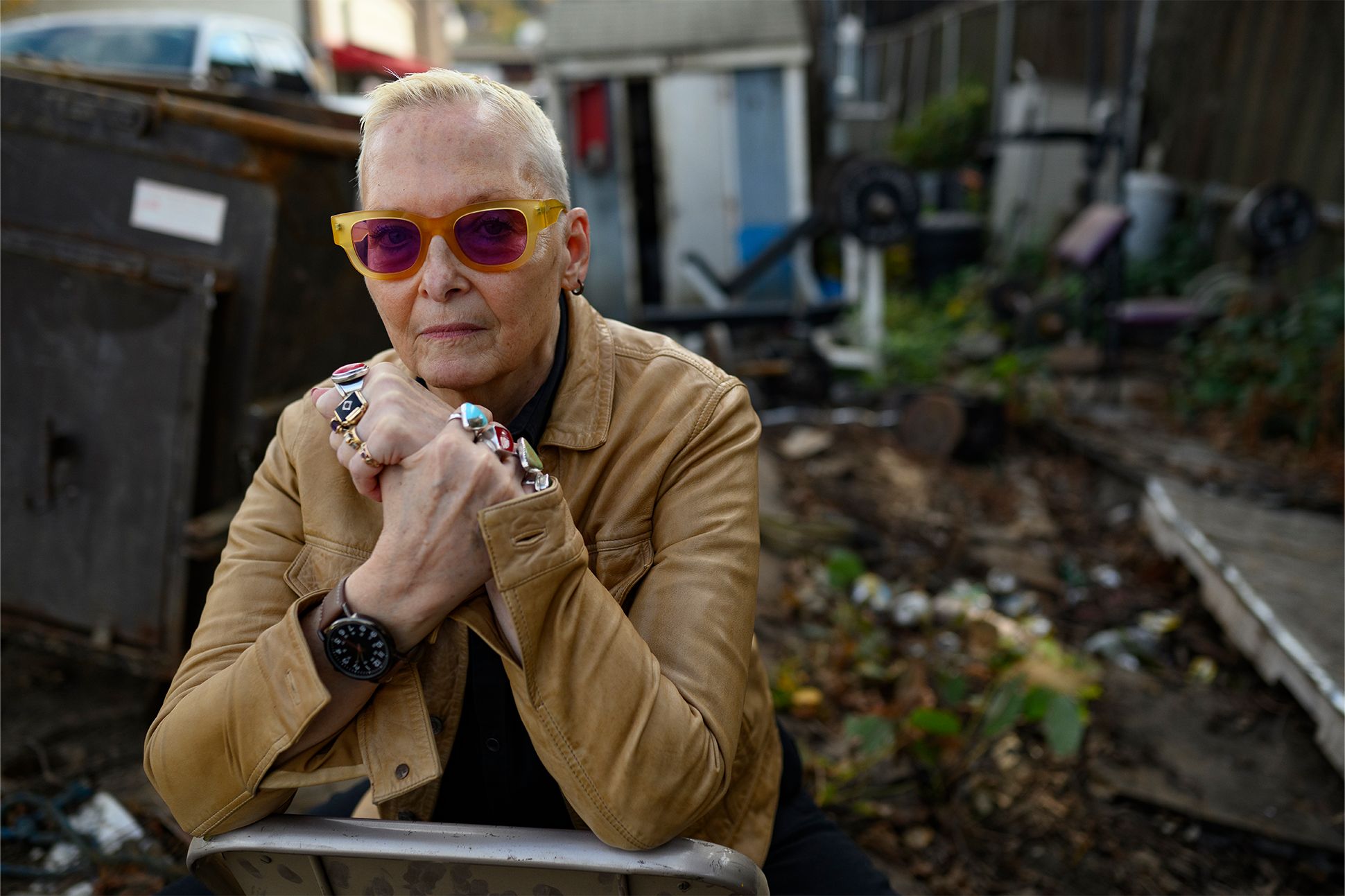 A photograph of Jan Beatty in sunglasses and a tan leather jacket, sitting in a chair facing the camera. Behind her is a dumpster and various littered objects.