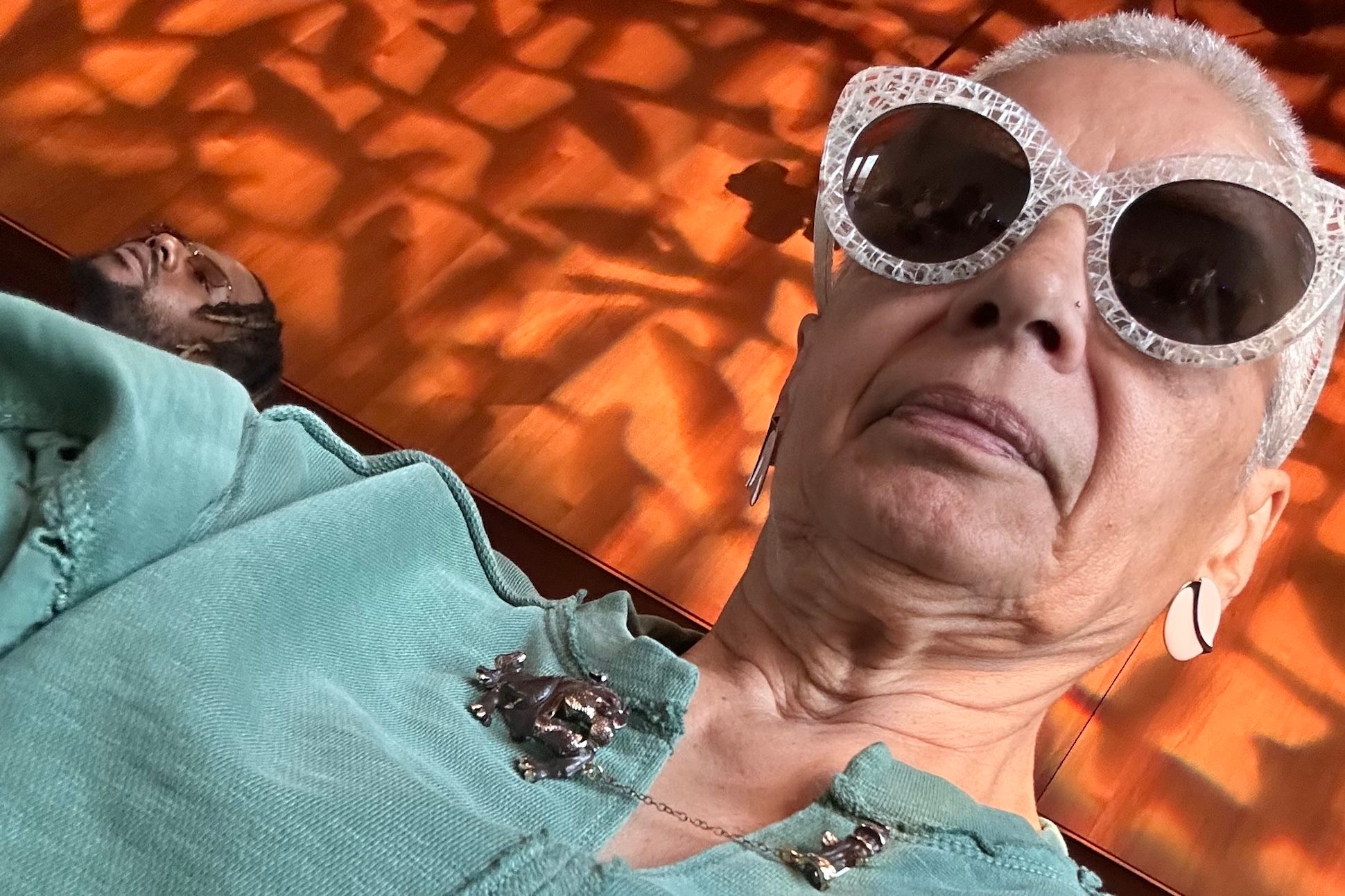 Marta Collazo, an Afro-Boricua woman with short cropped white hair, wearing a seafoam top with a
bulldog pin and big round white sunglasses, poses for a picture in front of a
cherry wood background.
