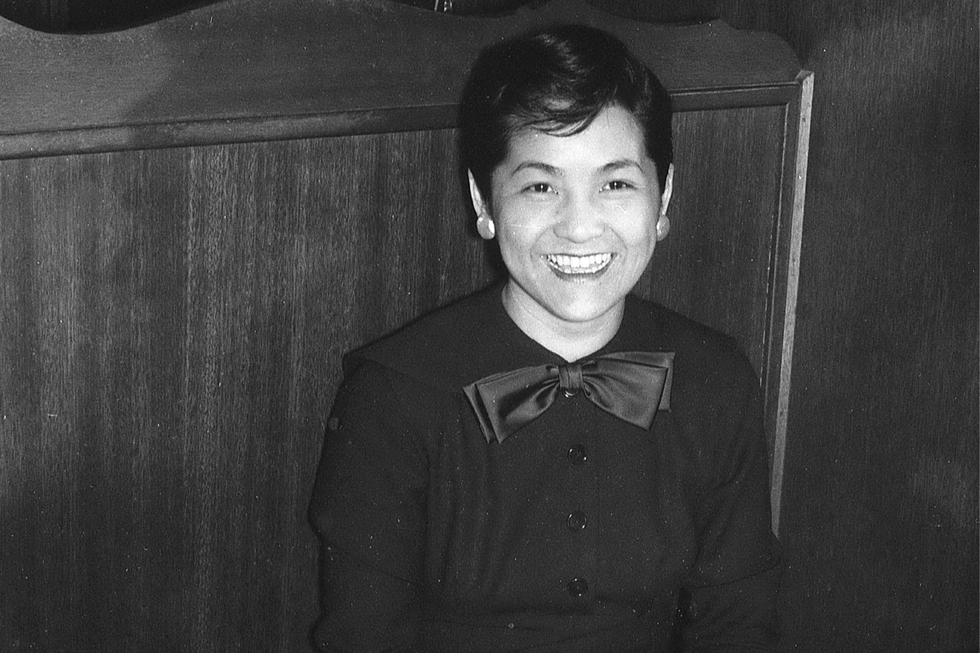 A black-and-white photograph of Nellie Wong, sitting in a booth, wearing a silk bow tie and a dress.