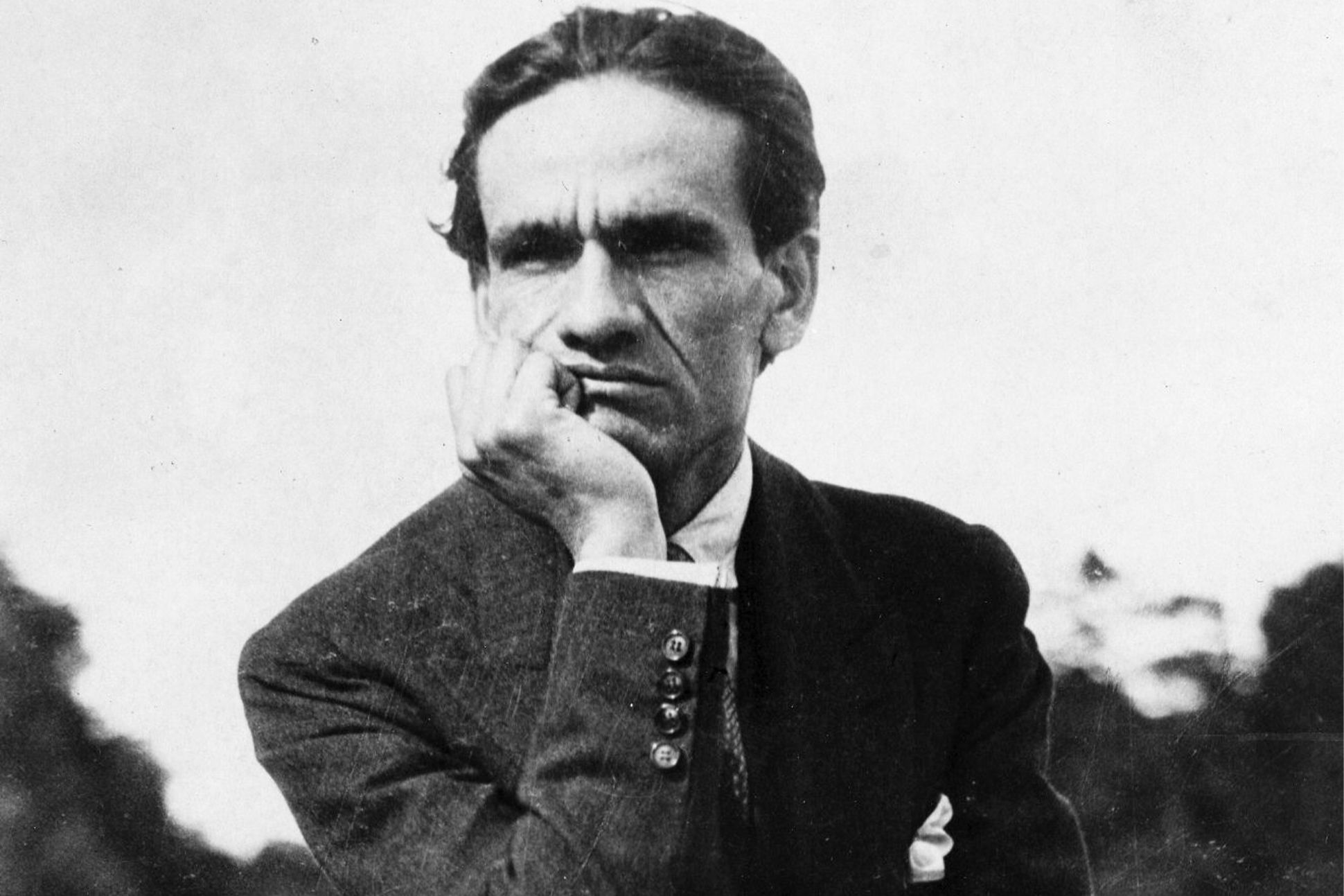 A black-and-white photograph of César Vallejo sitting outdoors, chin in hand, gazing off into the distance.