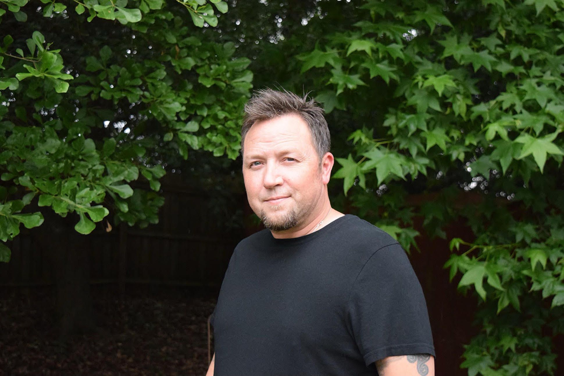 A white man smiling in a black t-shirt in front of a tree