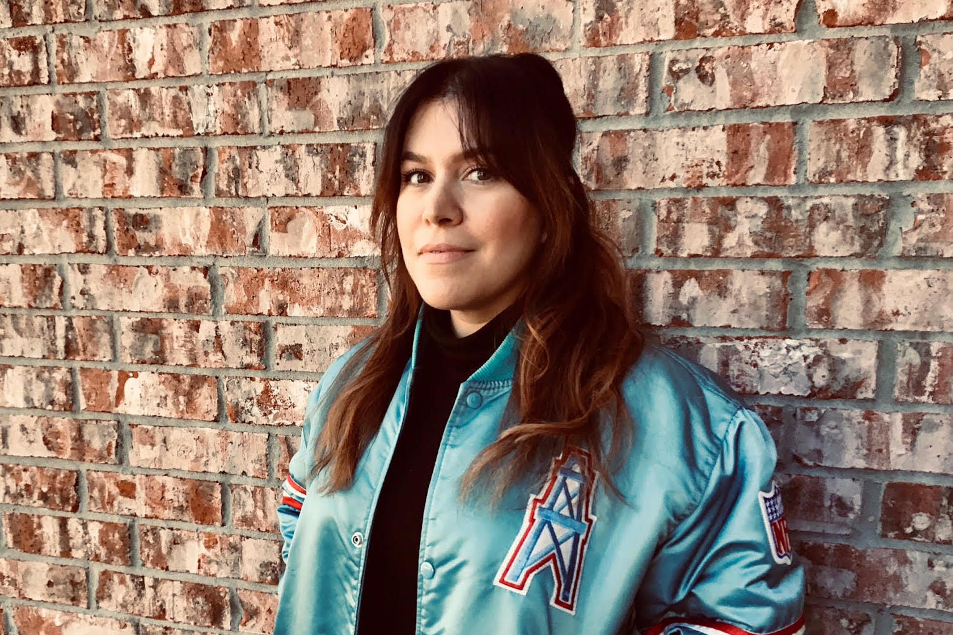 Poet Rocho Illiana standing in front of a brick wall, wearing a metallic blue bomber jacket.