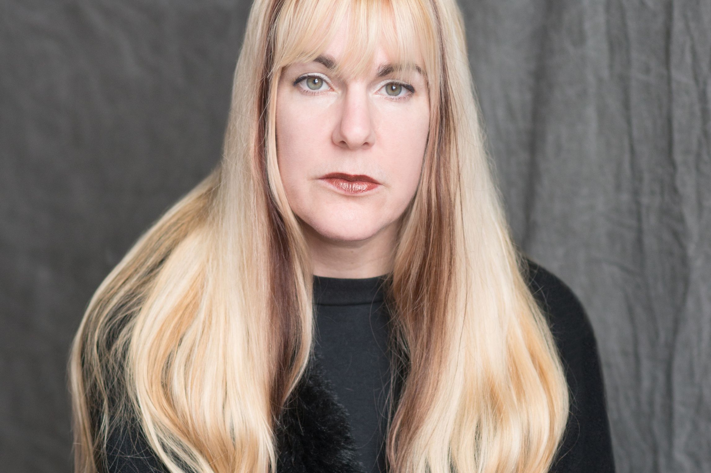 A white woman with long blond hair and a black blouse sitting in front of a grey background.