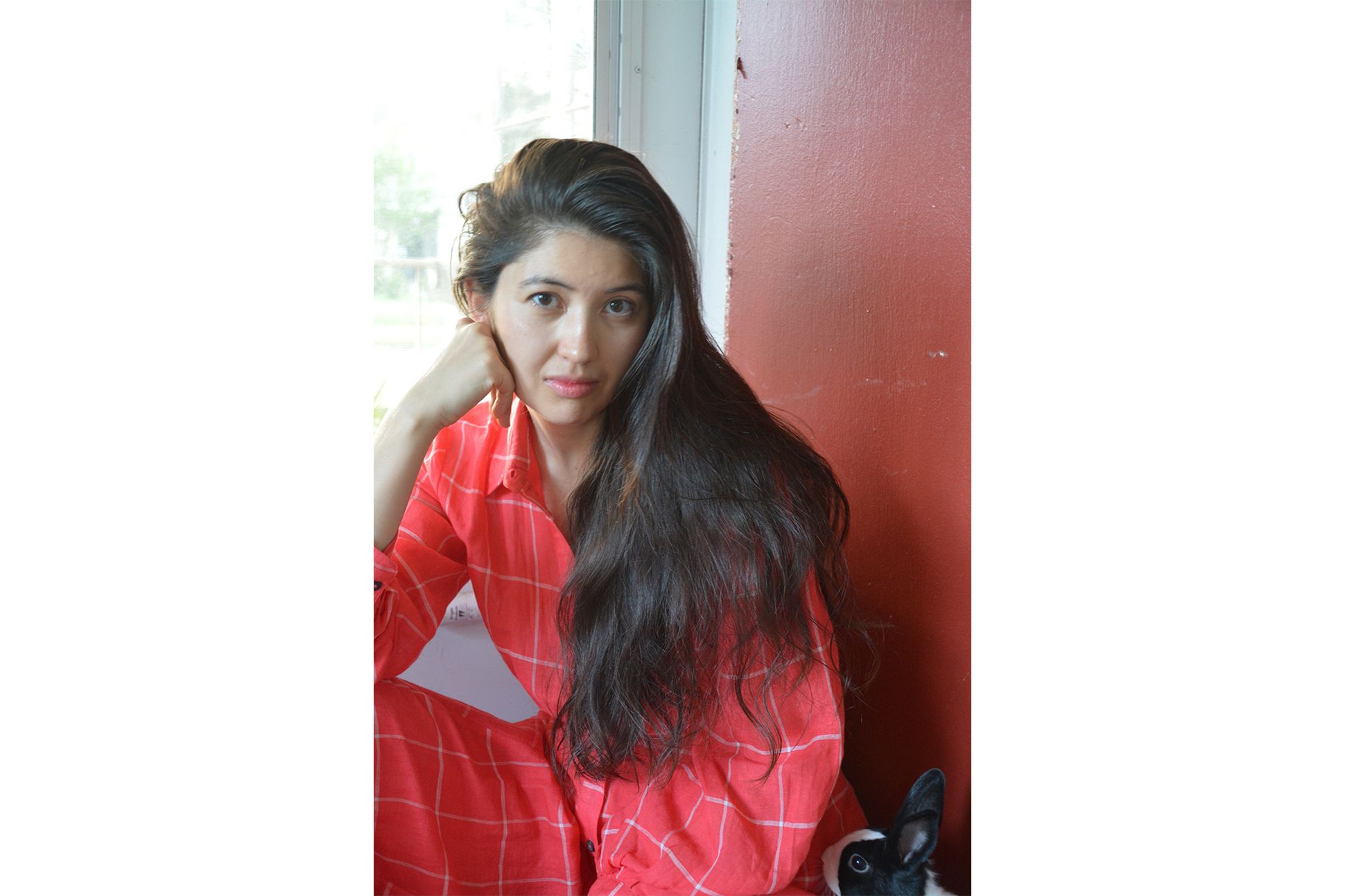 A person with long hair sits in front of a red door in a red dress.