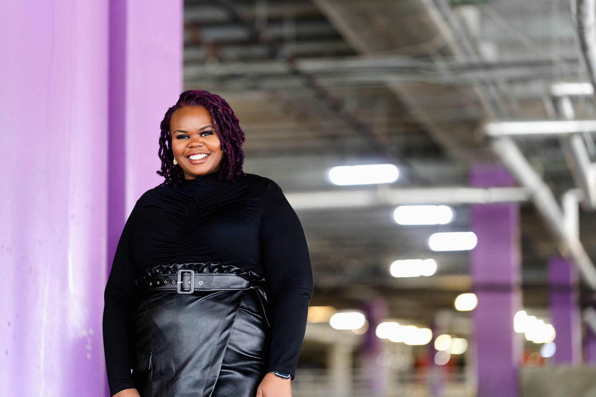 A black woman in a black pullover and leather skirt smiling next to a purple pillar