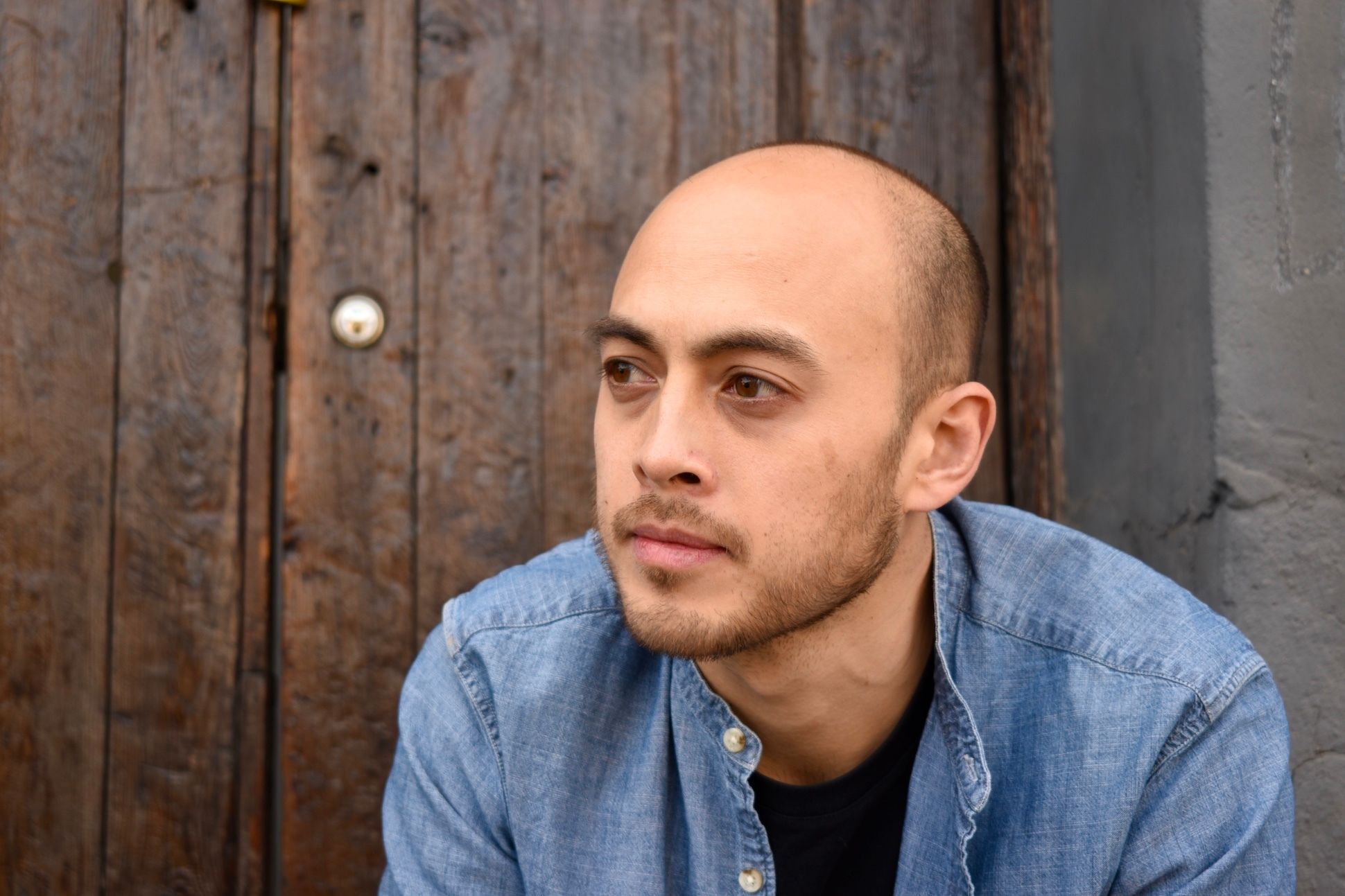 Man with skinhead hairstyle in a denim jacket sitting in front of a wooden door