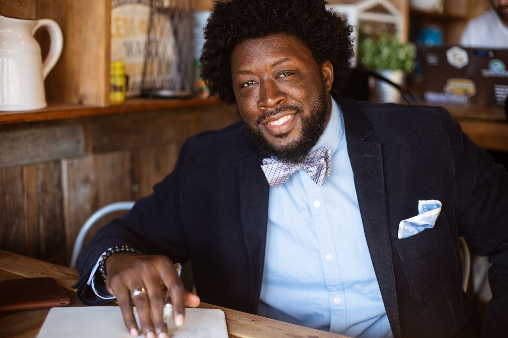 Black man in a suit with a bow tie sitting in a cafe and smiling with a pen and notebook