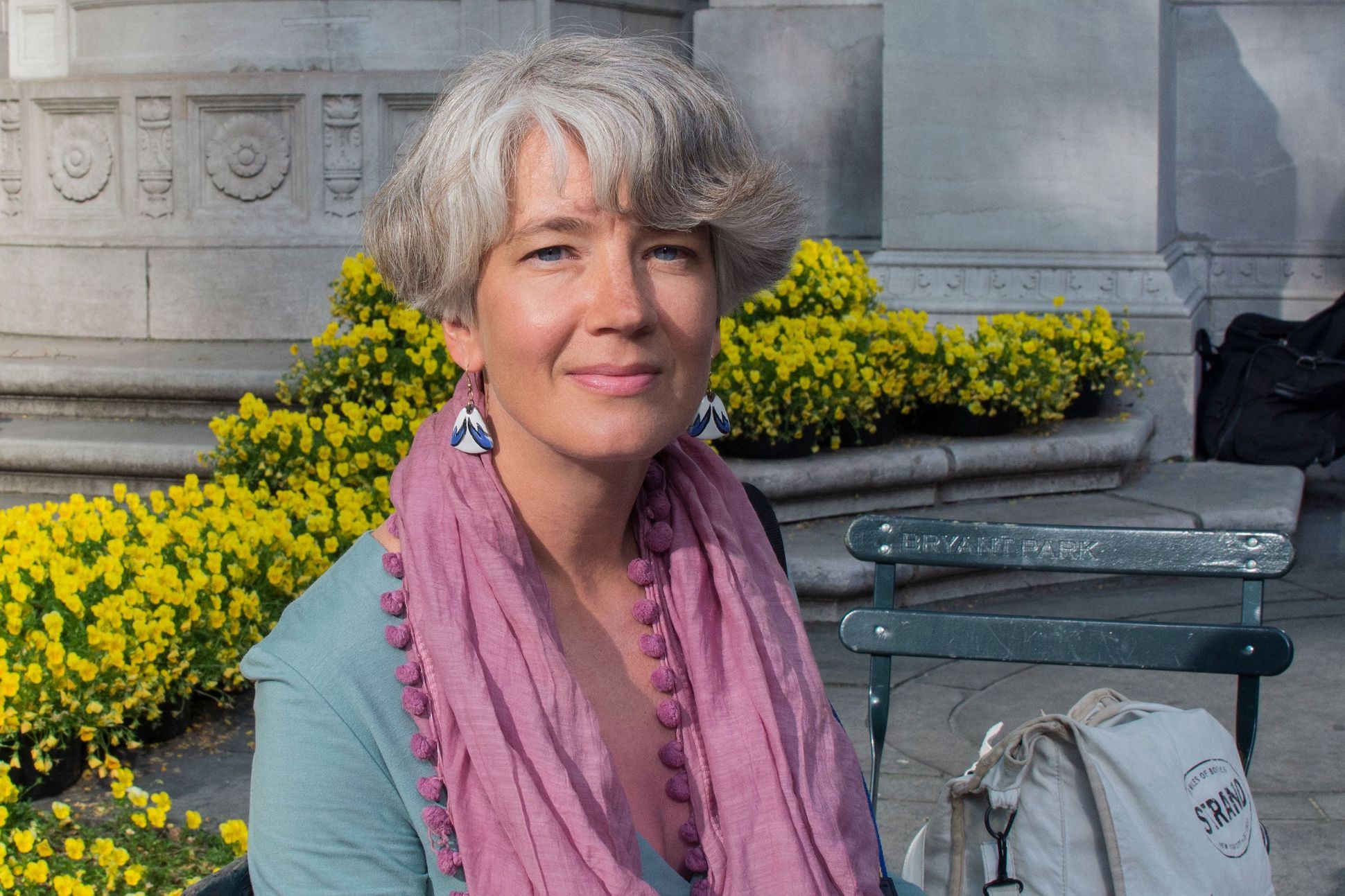 A woman with short grey hair sitting on a bench outside a historic building. She is wearing a powder pink scarf and turquoise blue