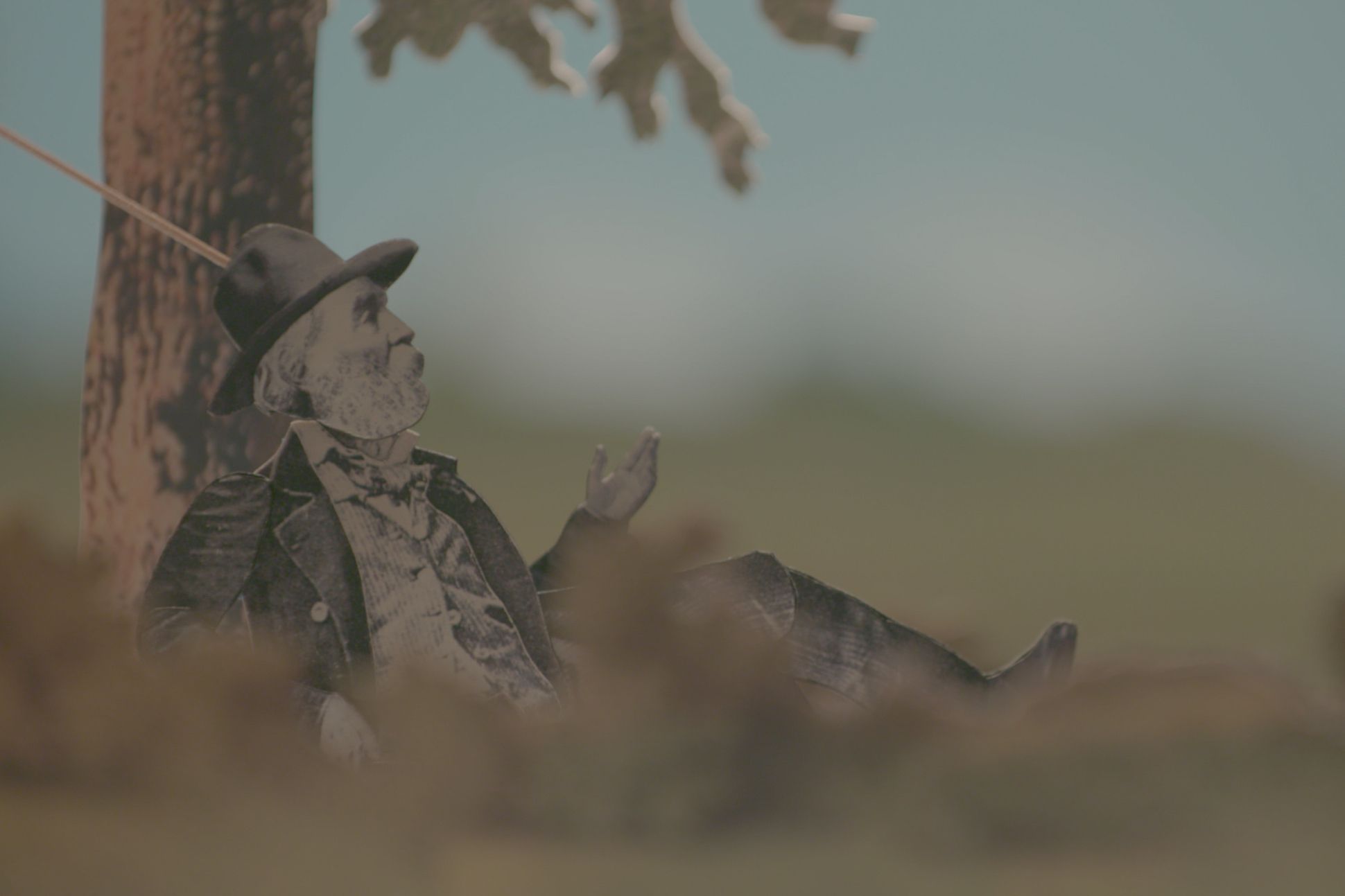 Blurry image of a black & white cut-out of Whitman leaning against a tree in a grassy meadow.