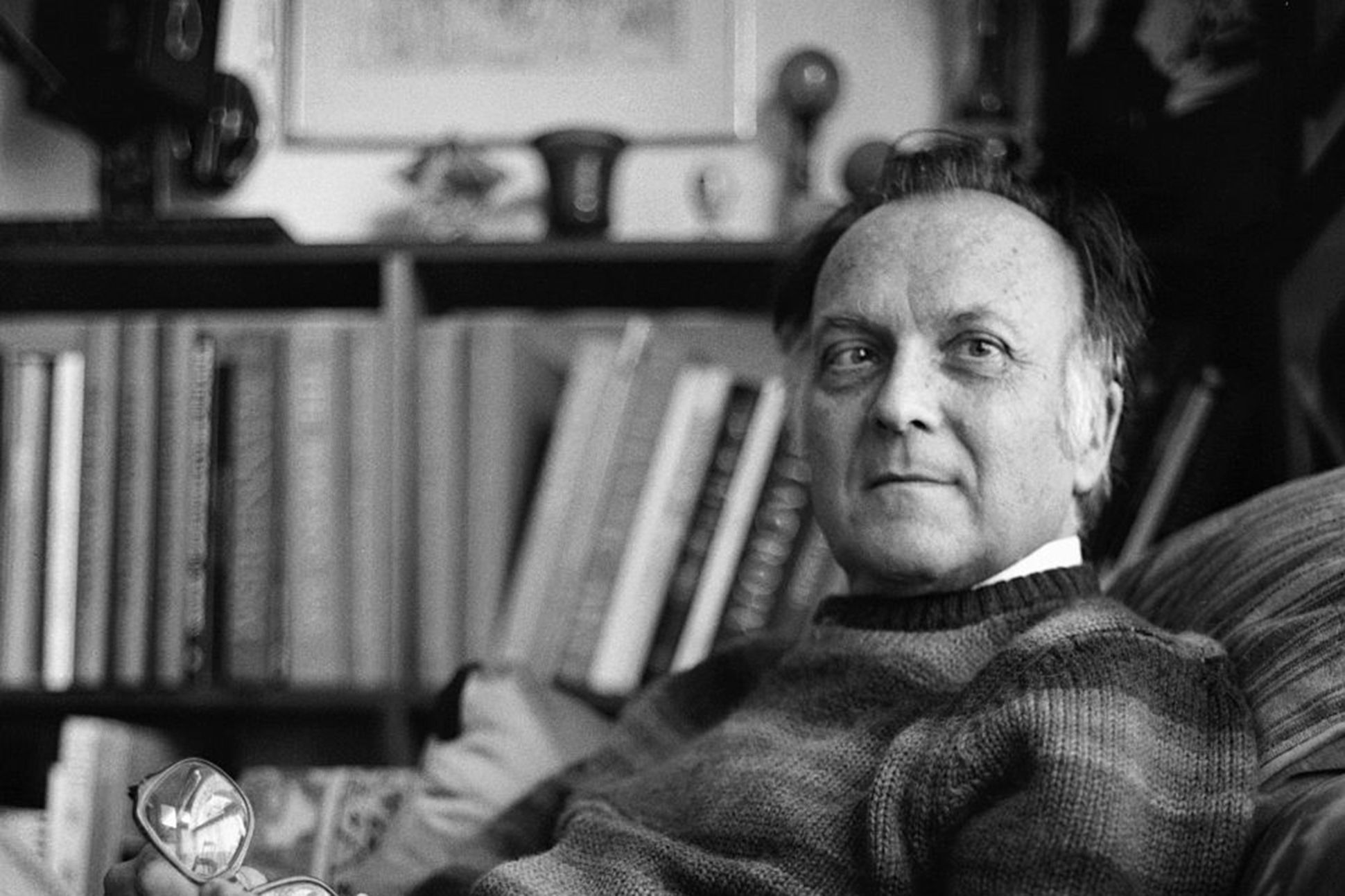 Black and white portrait of the poet Robert Duncan, reclining in an armchair in his study.