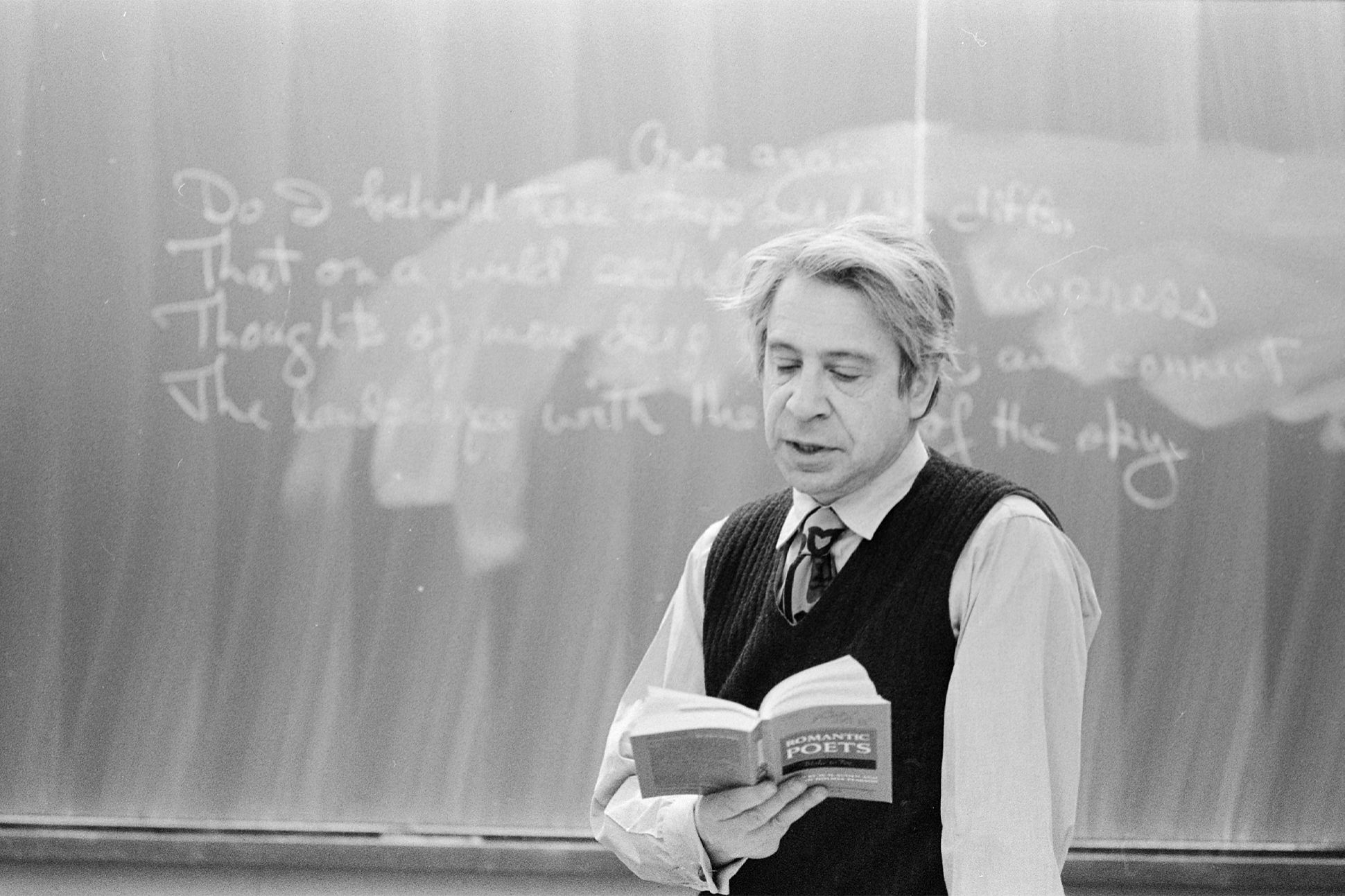 Black-and-white photo of Anthony Hecht standing in front of a blackboard, reading from a book of poetry.