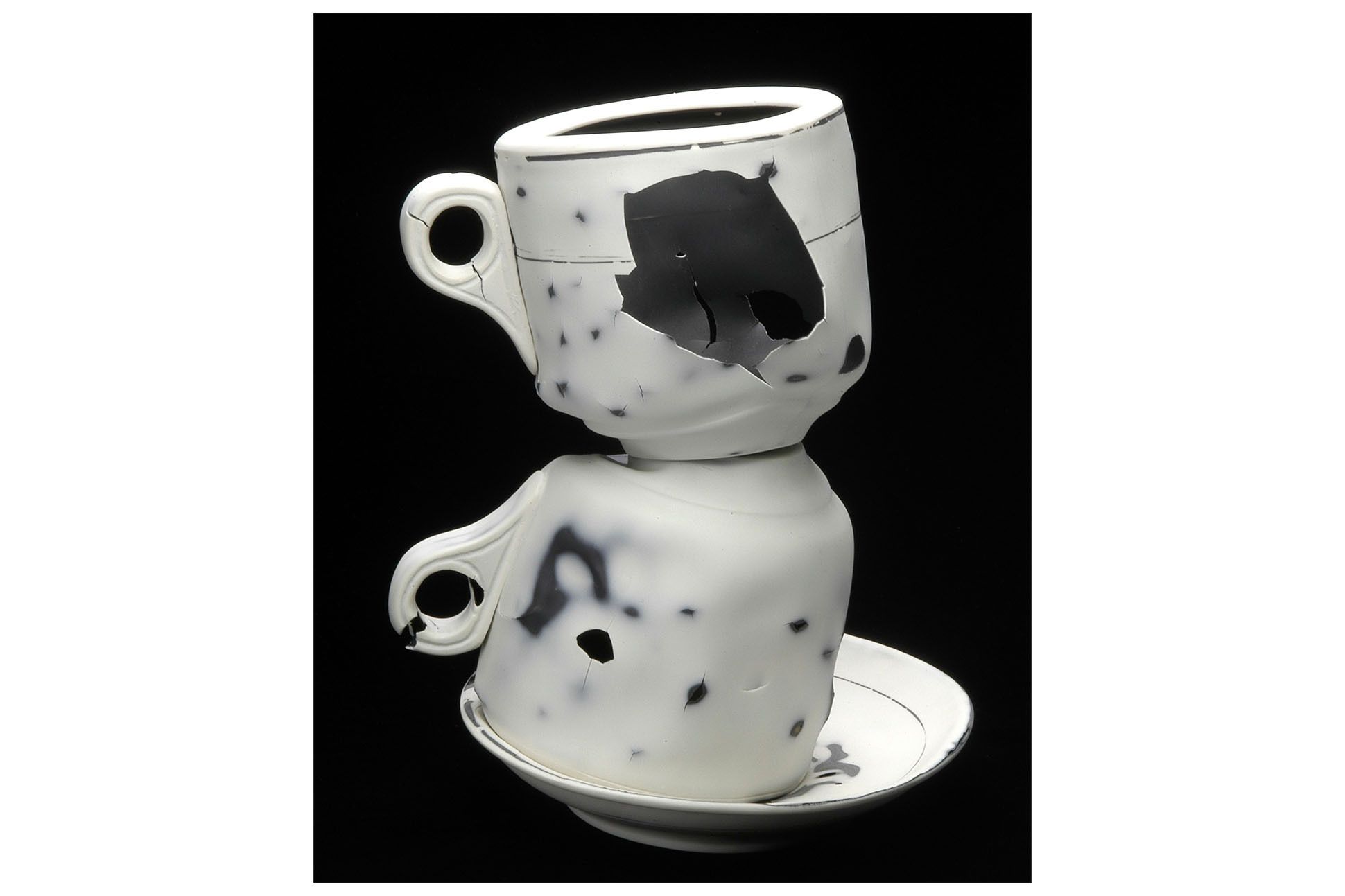 Photograph of art installation, two broken white teacups with stains and holes, piled on top of each other, in a saucer.