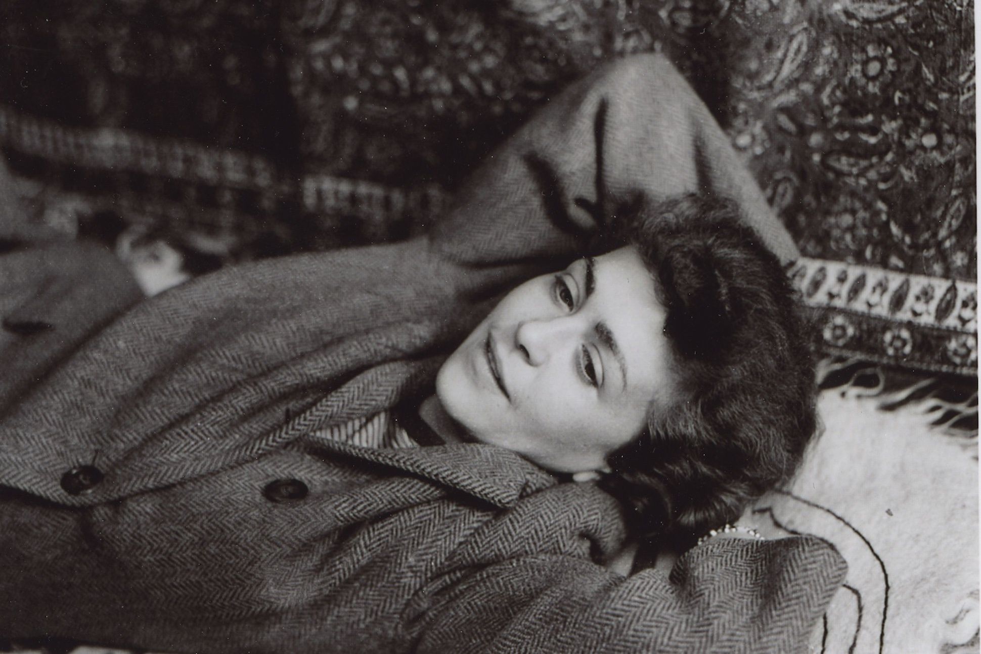 Black and white portrait of poet Jean Garrigue wearing a coat and lying on a rug with her hands on the back of her head