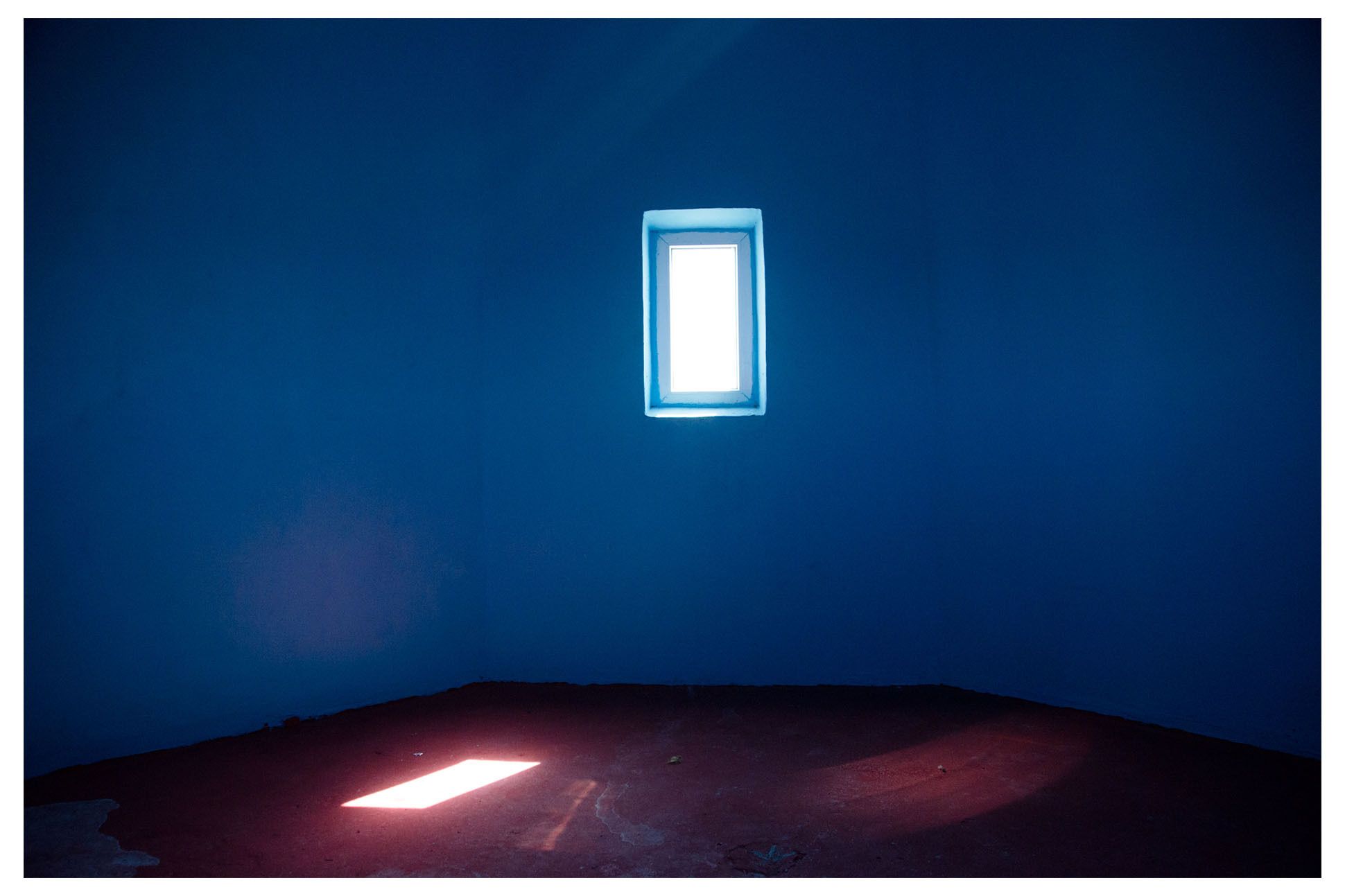Interior of a room with sun streaming in through a small rectangular window. Blue walls, dark floor,