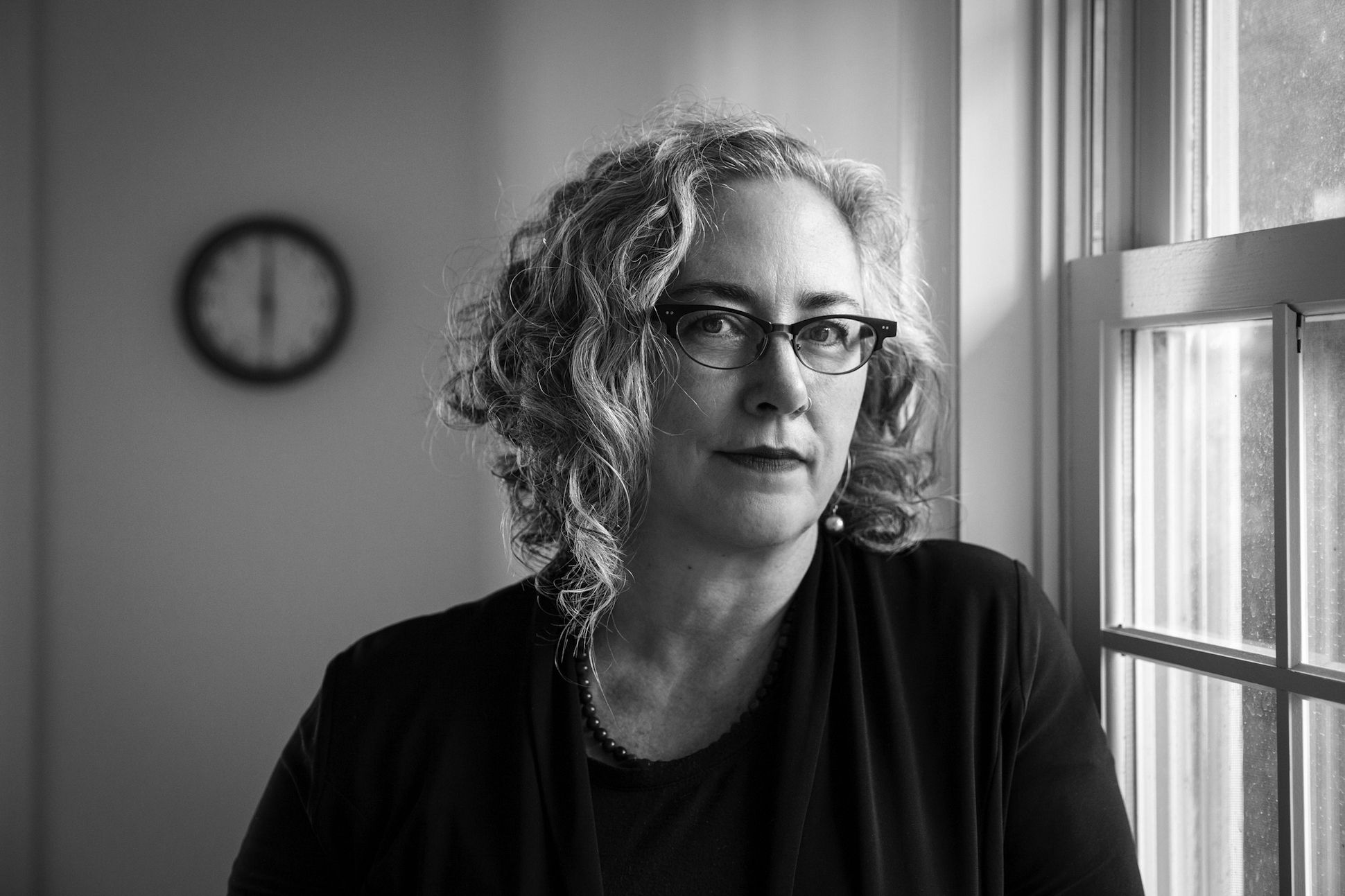 Black and white portrait of a woman wearing glasses and sitting beside a window in front of a clock