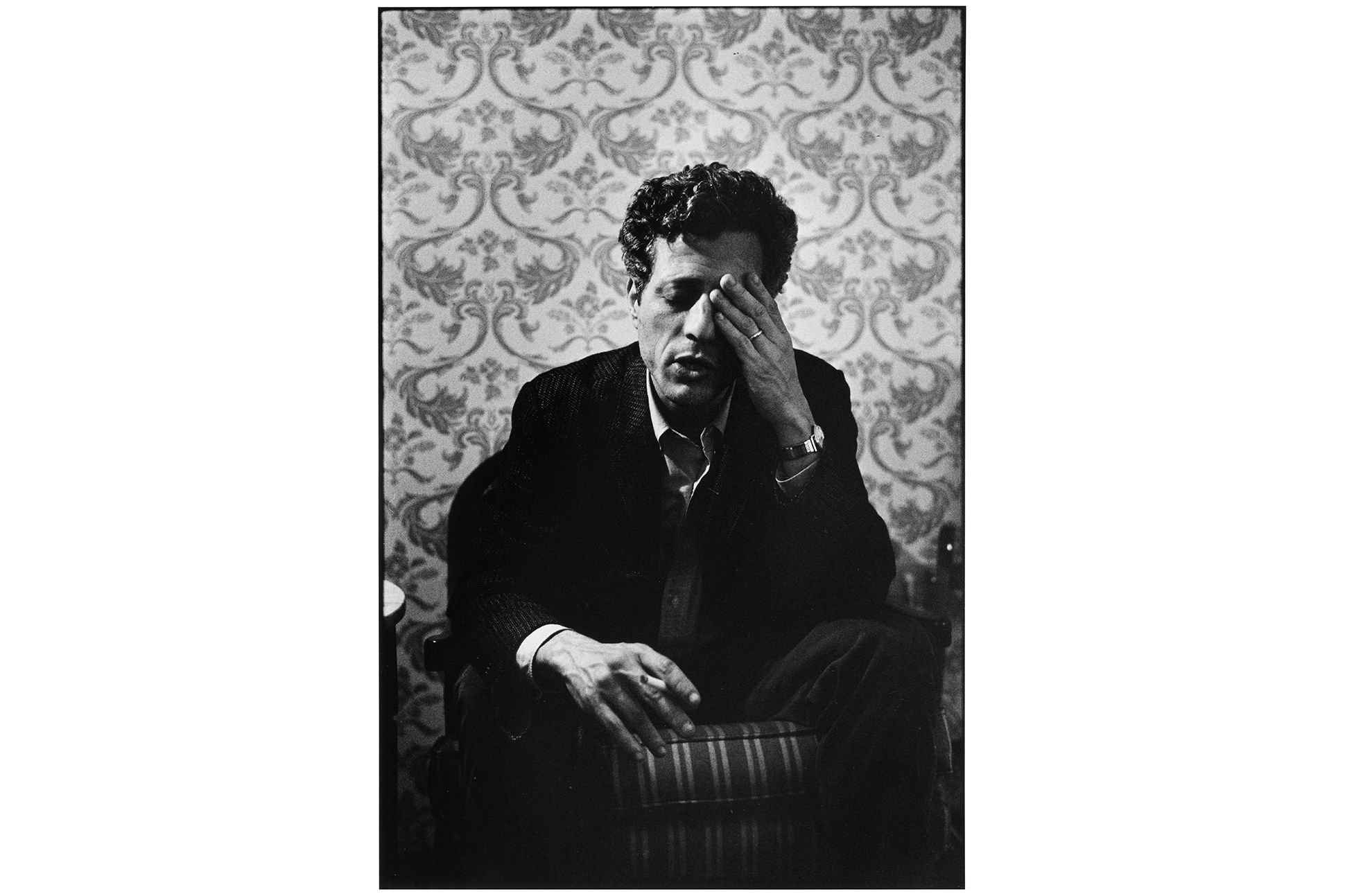 Man with a hand covering one eye sitting in front of wallpapers with floral pattern