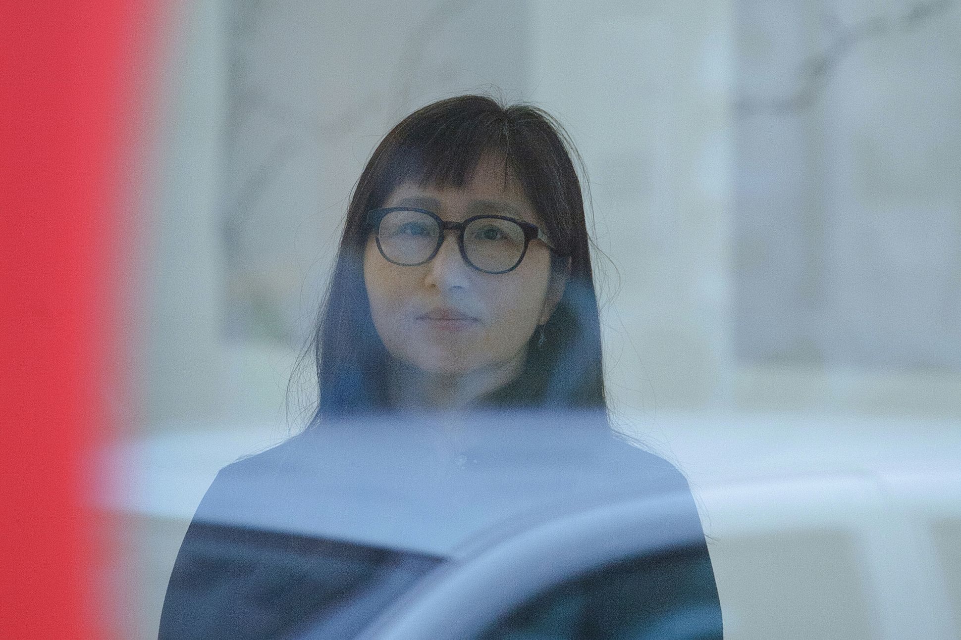 An Asian woman with long hair and glasses standing behind a glass panel