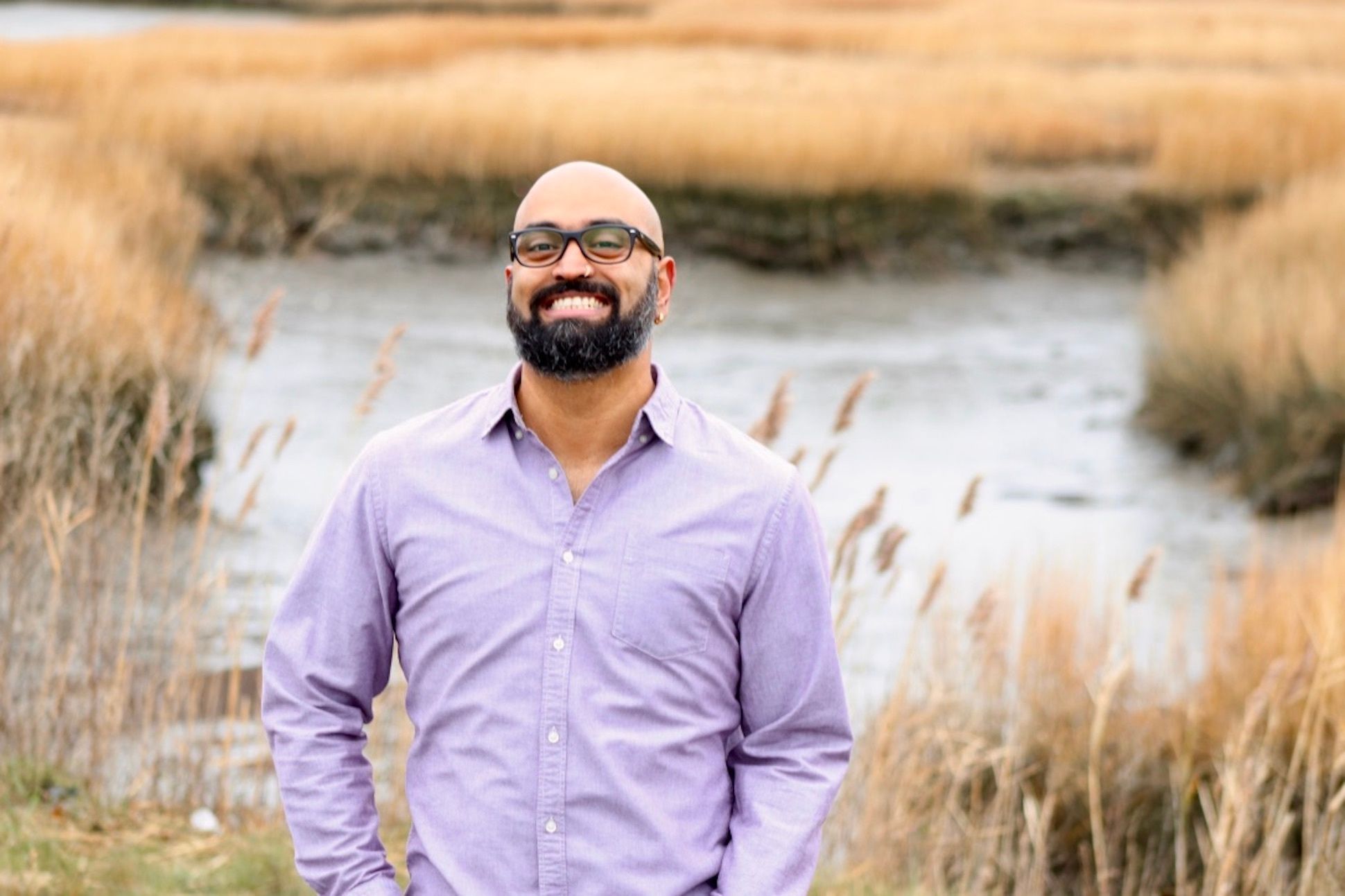 Bald man with beard and glasses wearing a purple shirt and standing in a cattail field.