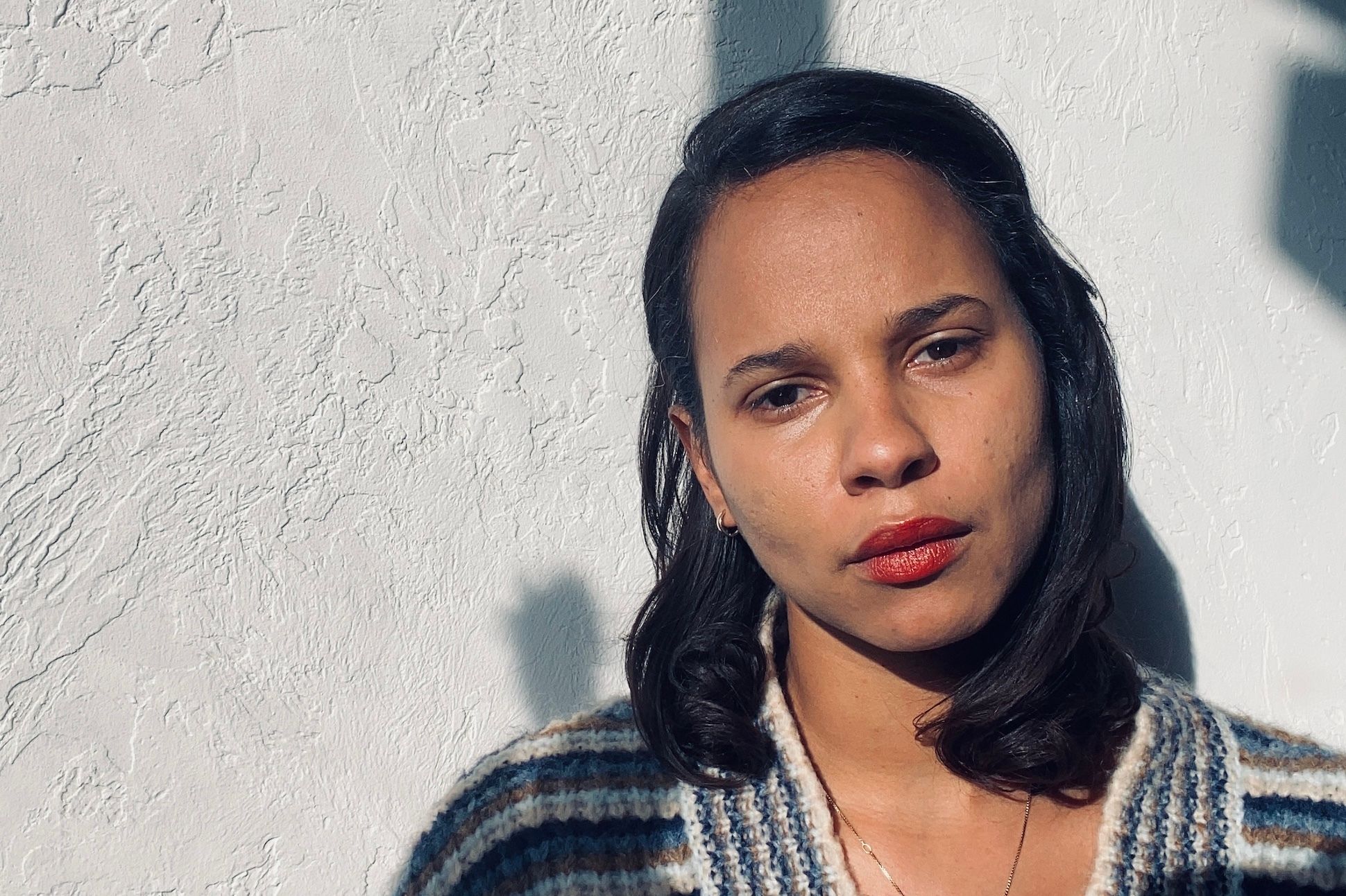 An African American woman with medium length hair tilting her hand in front of a white wall under the sun