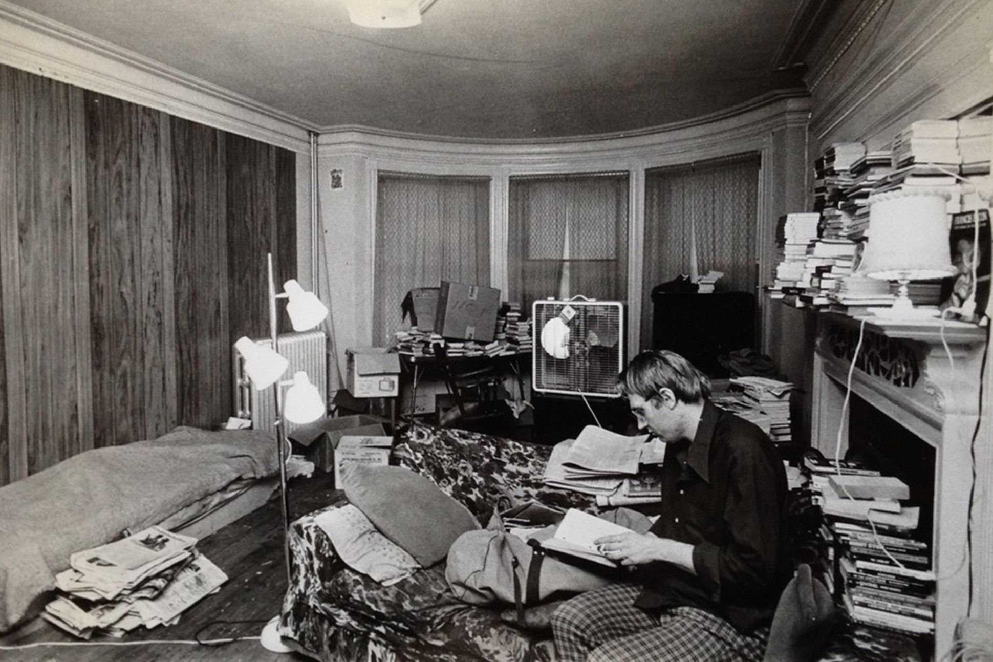 A black-and-white photograph of the poet Bill Knott reading a book in a messy room piled with books, newspapers, and furniture.
