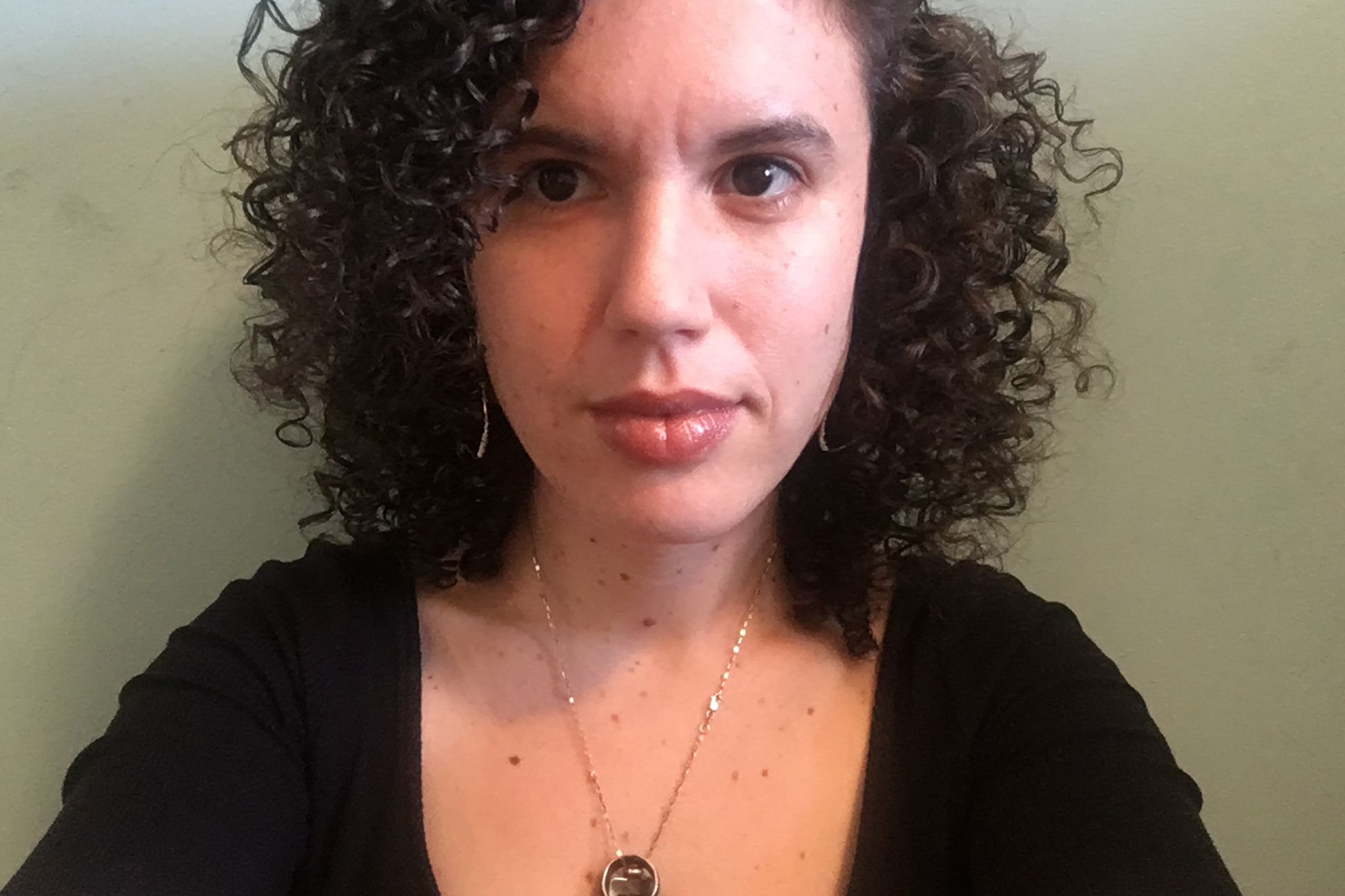 poet claire scwartz wearing a black shirt with a necklace standing in front of a wall.