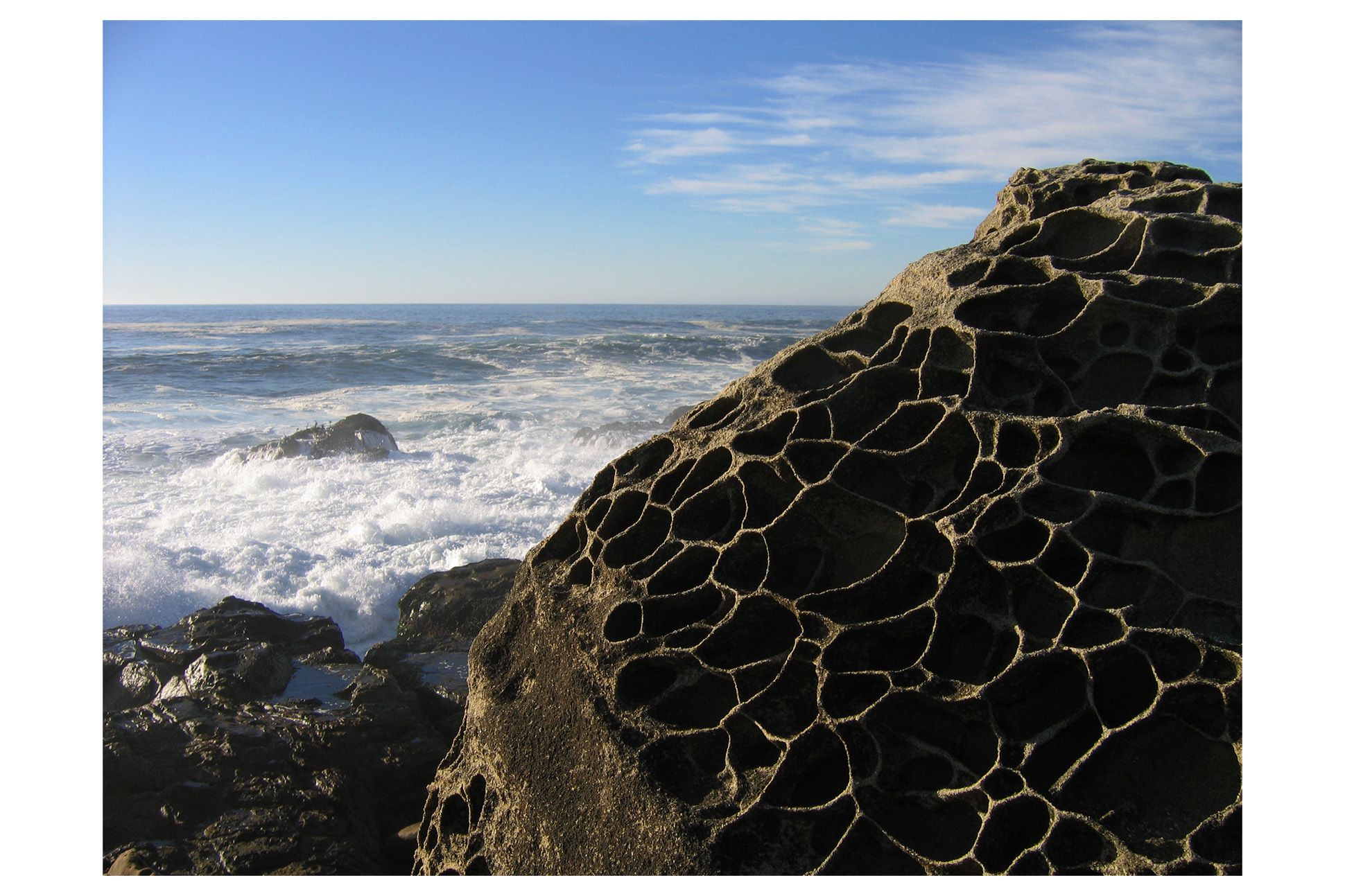 View of ocean and blue skies, with waves lapping up against rocks. Large Tafoni rock in foreground right.