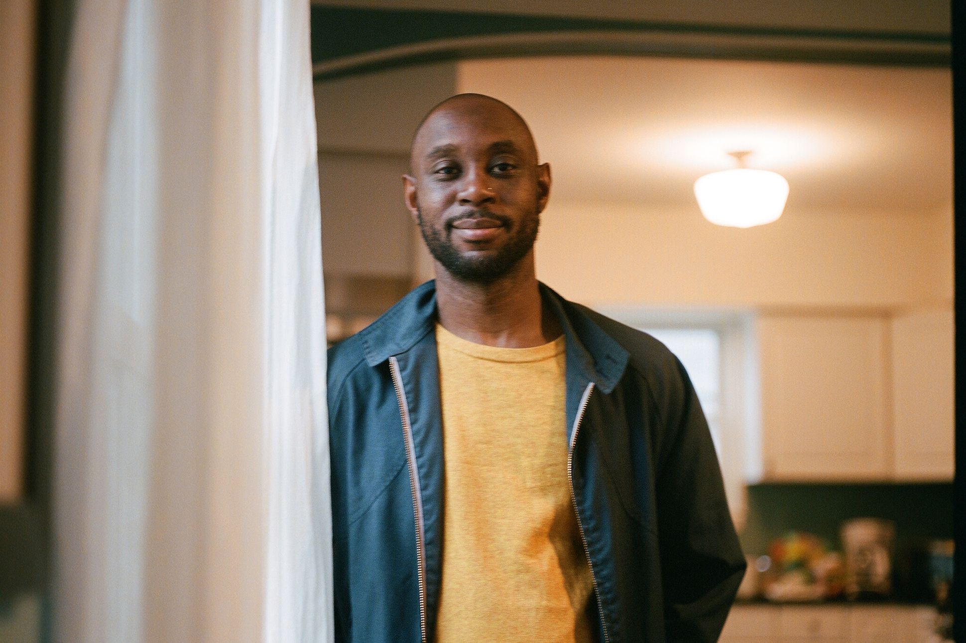 A black man in a yellow t-shirt and blue jacket standing by the window curtain