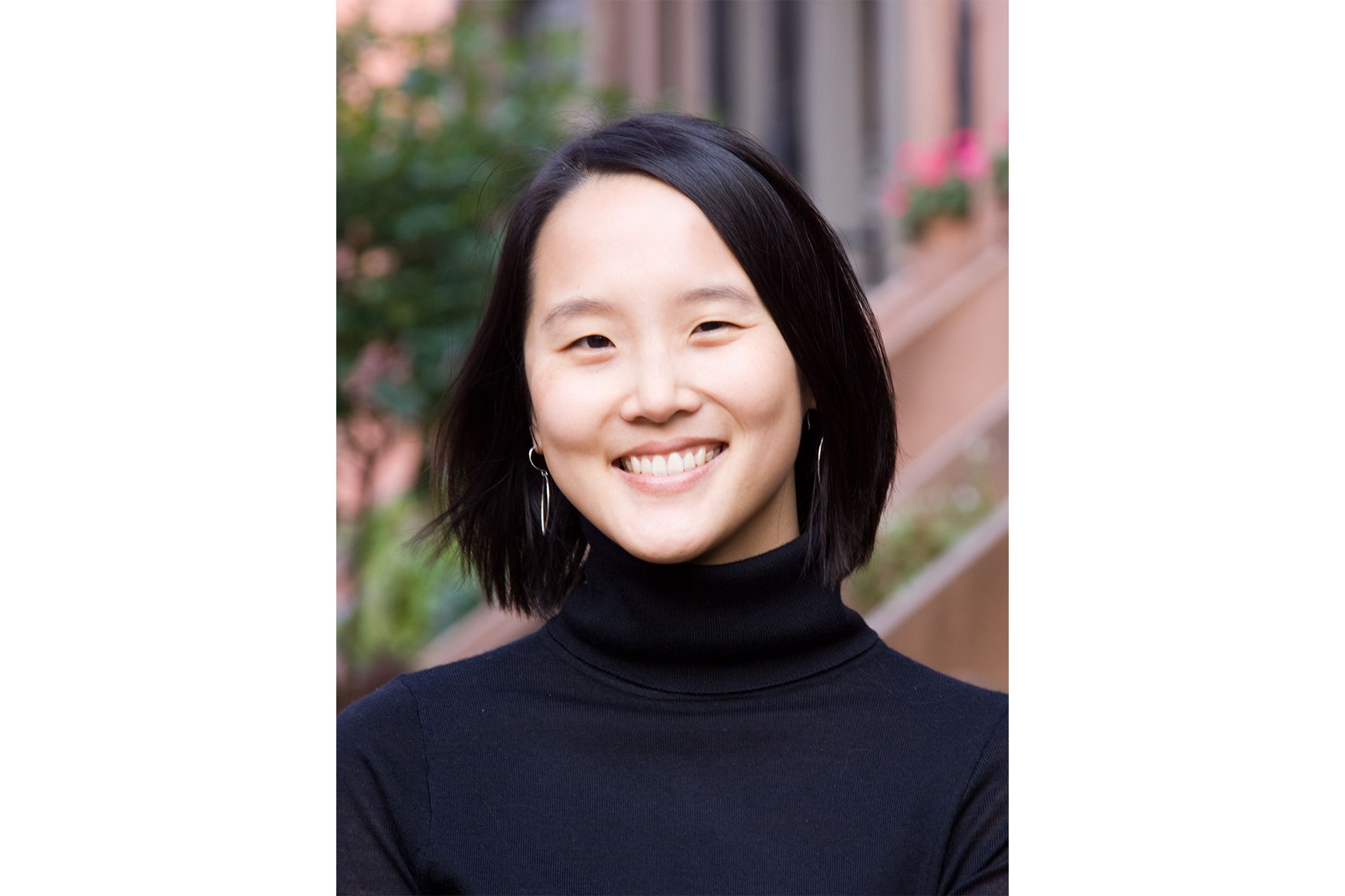 An Asian woman in a black turtleneck sweater smile in front of what appear to be the front porch of a house