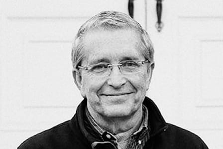 A white man in a jacket smiling in front of an entrance door