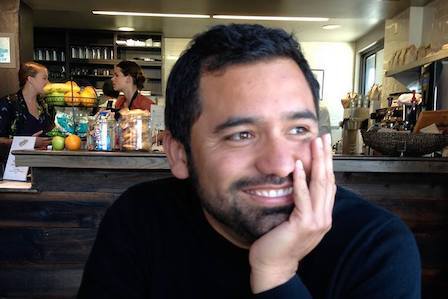 A hispanic man in a black jumper smiling with his hand on his jaw in a cafe