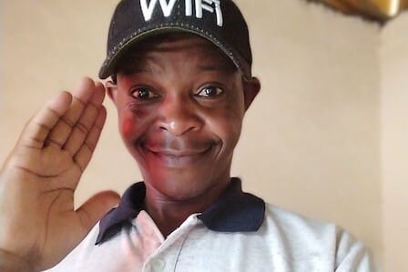 A black man with a cap and polo shirt waving at the camera