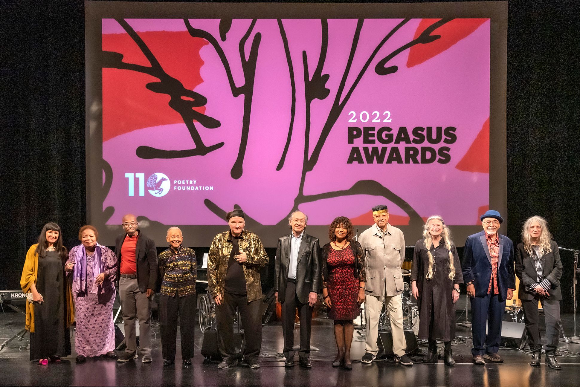 All standing on a stage in front of a pink and red projection that says "2022 Pegasus Awards" with illustrated pegasus (left to right): Sandra Cisneros, Angela Jackson, Kevin Quashie, Nikki Giovanni, CAConrad, Arthur Sze, Rita Dove, Haki Madhubuti, Sharon