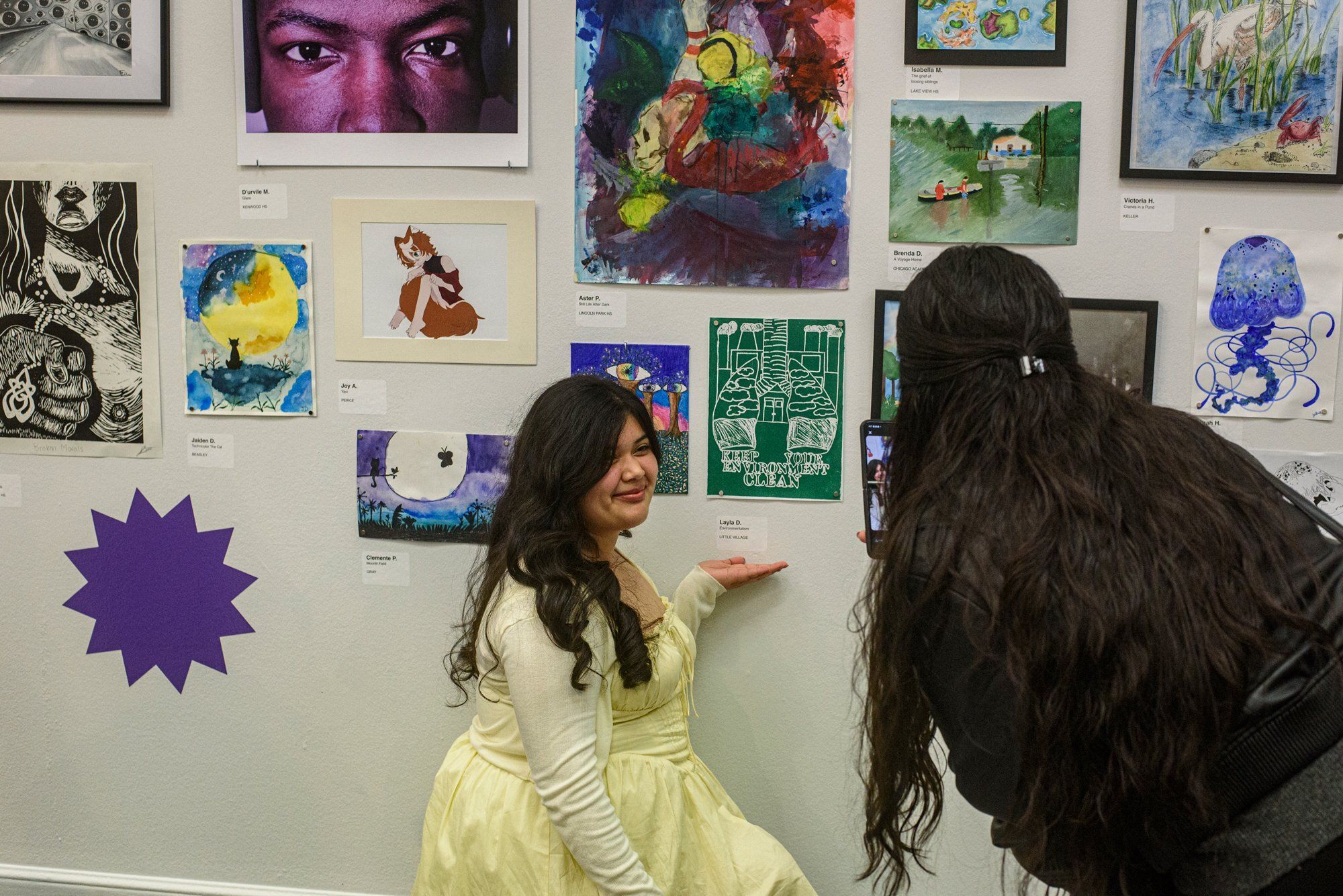 Student artist Layla D. poses with her artwork during the CPS All-City Visual Arts Exhibition at the Design Museum of Chicago. Layla is kneeling by a wall full of posters while someone bends down to look at it.